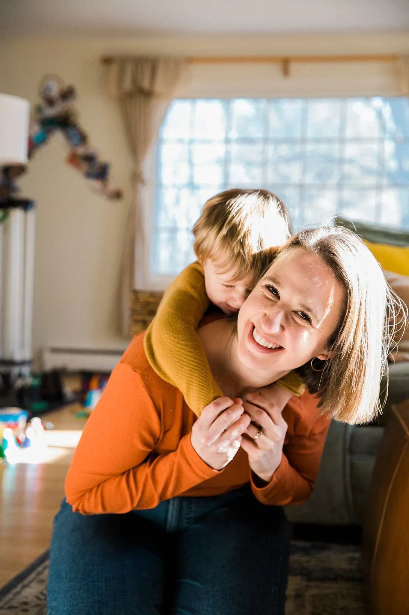 A woman with shoulder-length blonde hair and earrings smiling while holding a young boy on her back. The boy has blonde hair and is leaning in close to her, smiling. They are in a cozy room with a window and sunlight coming in.