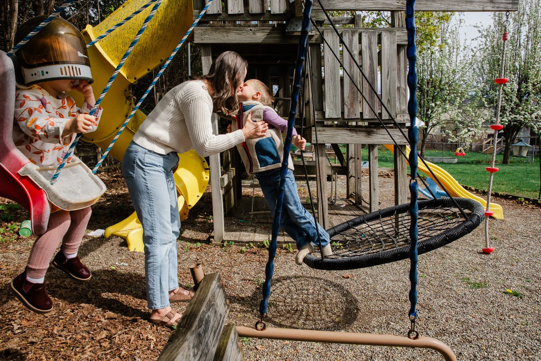 A woman and two children on a playground. The woman is playing with a young boy, possibly a sibling, while a girl is on a swing nearby. The playground has a wooden playhouse, a slide, and climbing ropes. The background includes green trees and a fenc