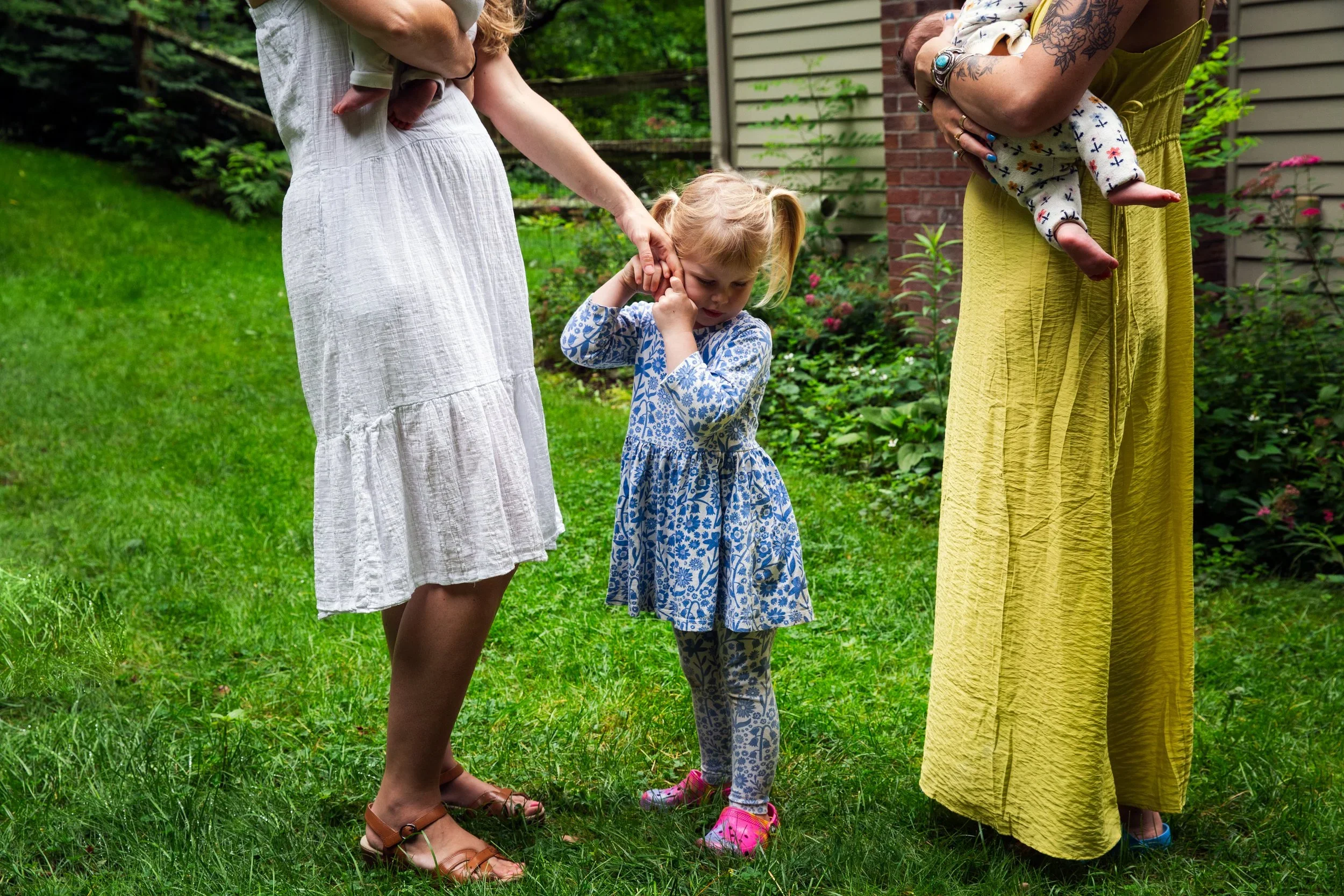 Two women and a young girl standing on a lush green lawn in front of a house with garden plants. One woman in a white dress is holding a baby, and the other woman in a yellow skirt is holding a small child near her chest, all engaged in a shared acti