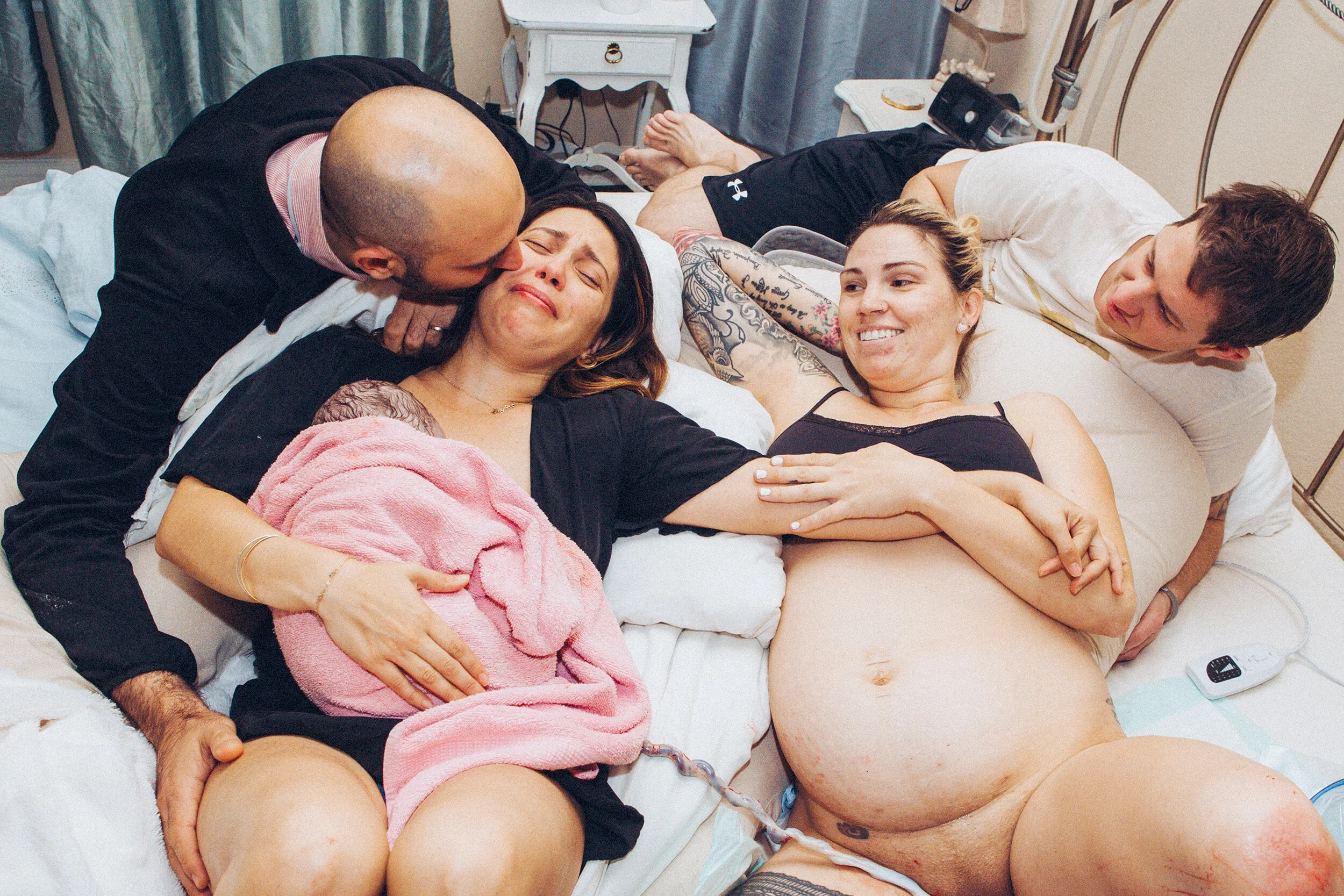 Two women and two men on a hospital bed, with one woman in labor holding her newborn, surrounded by friends or family. The father is kissing the mother, who is smiling and holding the baby. The scene is intimate and joyful.