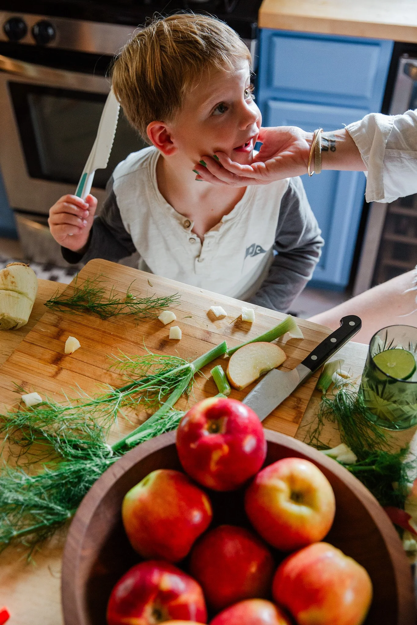A young boy with light brown hair and wearing a cream and gray shirt, sitting at a kitchen table with a wooden cutting board, chopping apples and garnishing with fresh herbs. An adult's hand supports his chin, and apples are in the foreground.