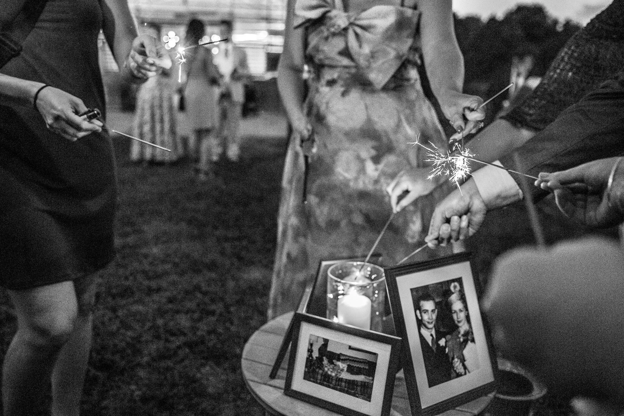 People holding sparklers at a gathering, with framed photos and candles on a table, likely celebrating an event or special occasion. The environment appears to be indoors or in a covered space.
