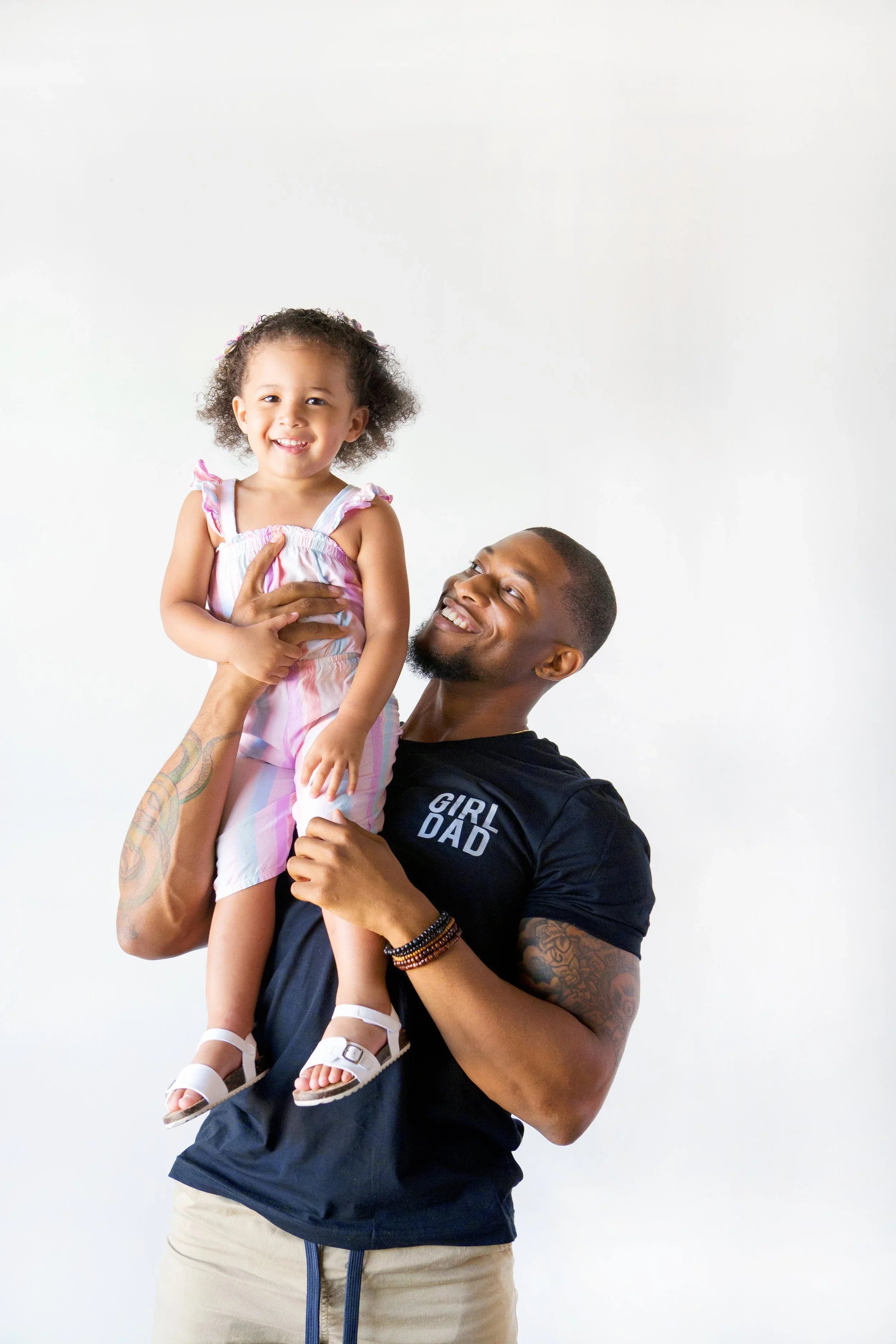 A young girl with curly hair, wearing a pastel pink and white dress with ruffled straps, is smiling and being held up by a man with a beard and tattoos on his arm. The man is wearing a black T-shirt with the text "GIRL DAD" on it. They are standing a