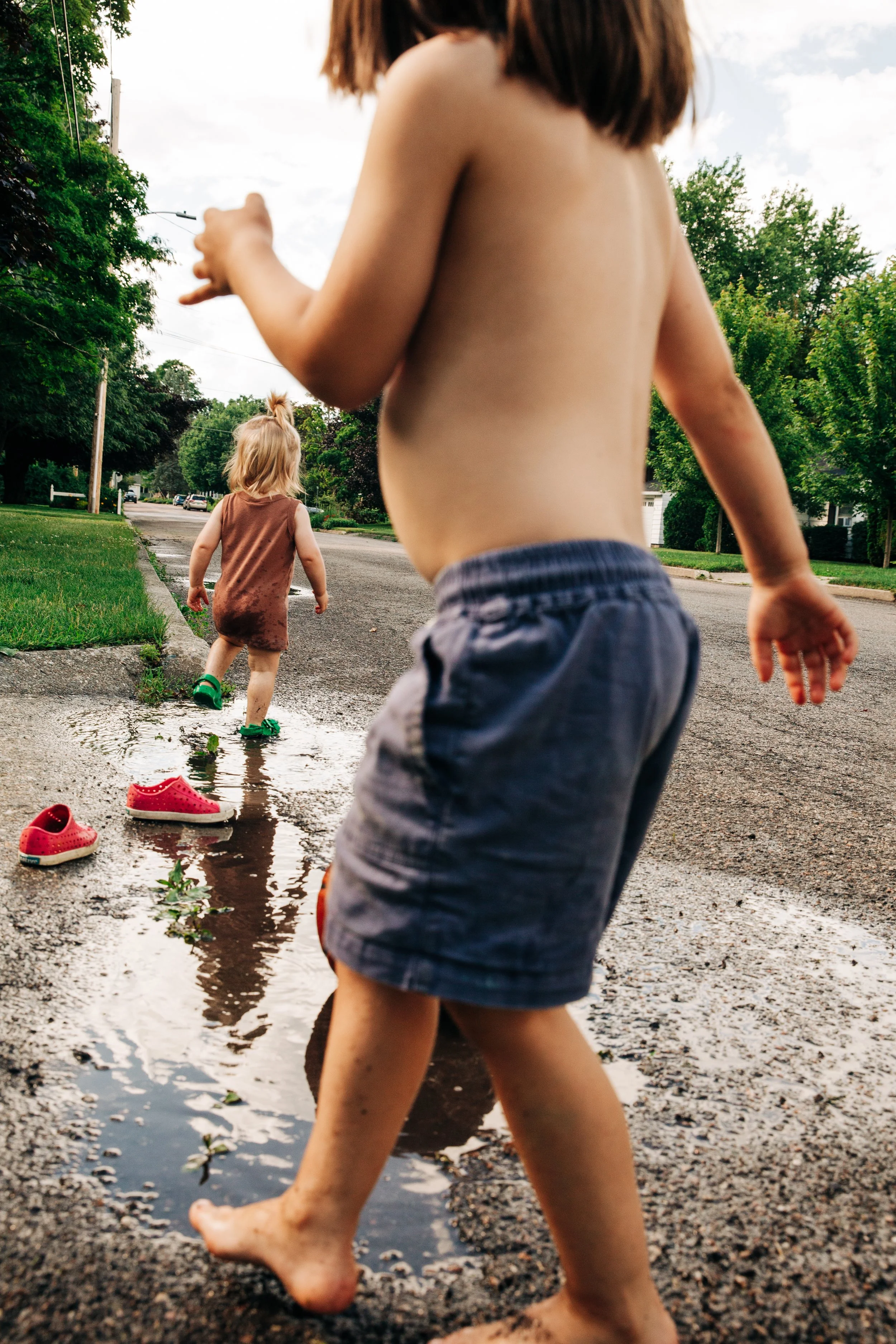Two children playing barefoot in a puddle on a residential street with green trees and partly cloudy sky.