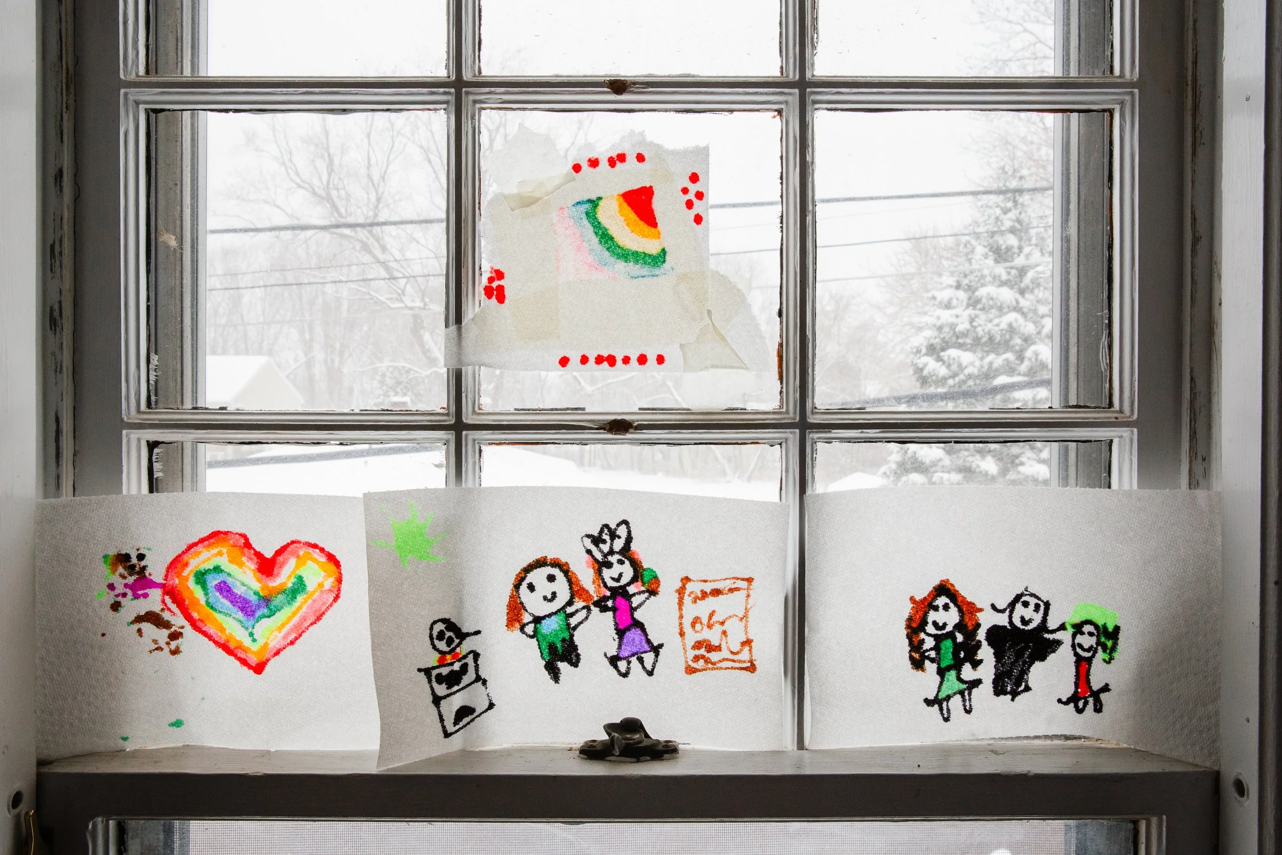 Children's drawings on paper taped to a window, with snowy outdoor scene visible through the glass.