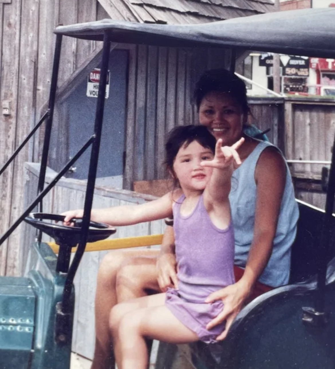 A woman and a young girl sitting on a golf cart, with the girl pointing and smiling at the camera, in front of a wooden fence.