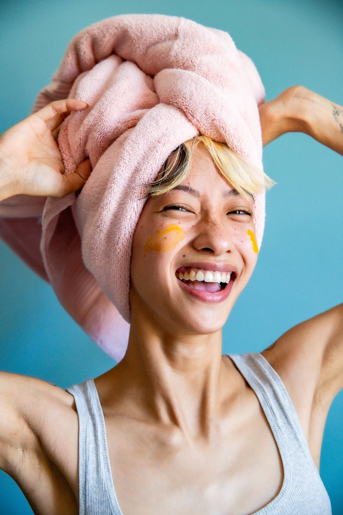 A young woman with short blonde hair smiling with her eyes closed, wrapped in a pink towel on her head, wearing a tank top, with yellow face masks on her cheeks, in front of a blue background.
