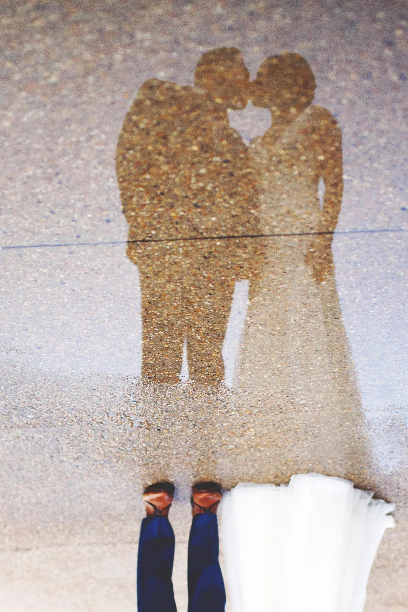 Reflection of a couple kissing on the ground, with the person taking the photo wearing brown shoes and blue pants, and the woman in a white dress with a white purse.