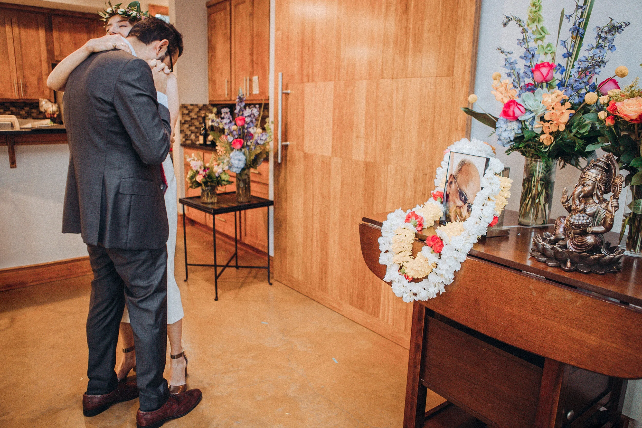 A couple is standing close together, embracing in a warm hug at a funeral or memorial service. They are near a table with floral arrangements, a photograph of the deceased surrounded by flowers, a statue of Ganesha, and various flower vases.