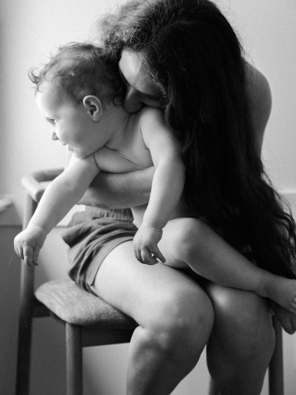 A black and white photo of a woman holding a young child on her lap, with the woman kissing the child on the head, both appearing to be relaxed and intimate.