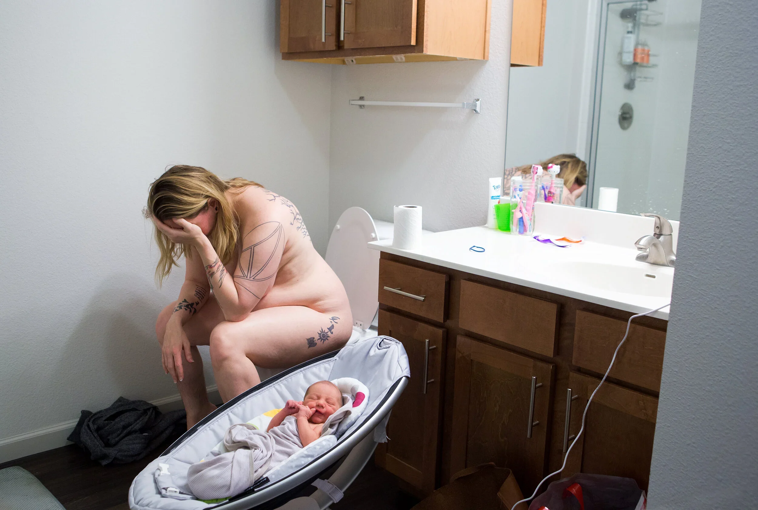 A woman with tattoos on her arms and chest, sitting on the toilet in a bathroom, holding her head in her hand and looking down, with a newborn baby lying in a bassinet nearby. The bathroom has a white wall, wooden cabinets, a sink with toiletries, an