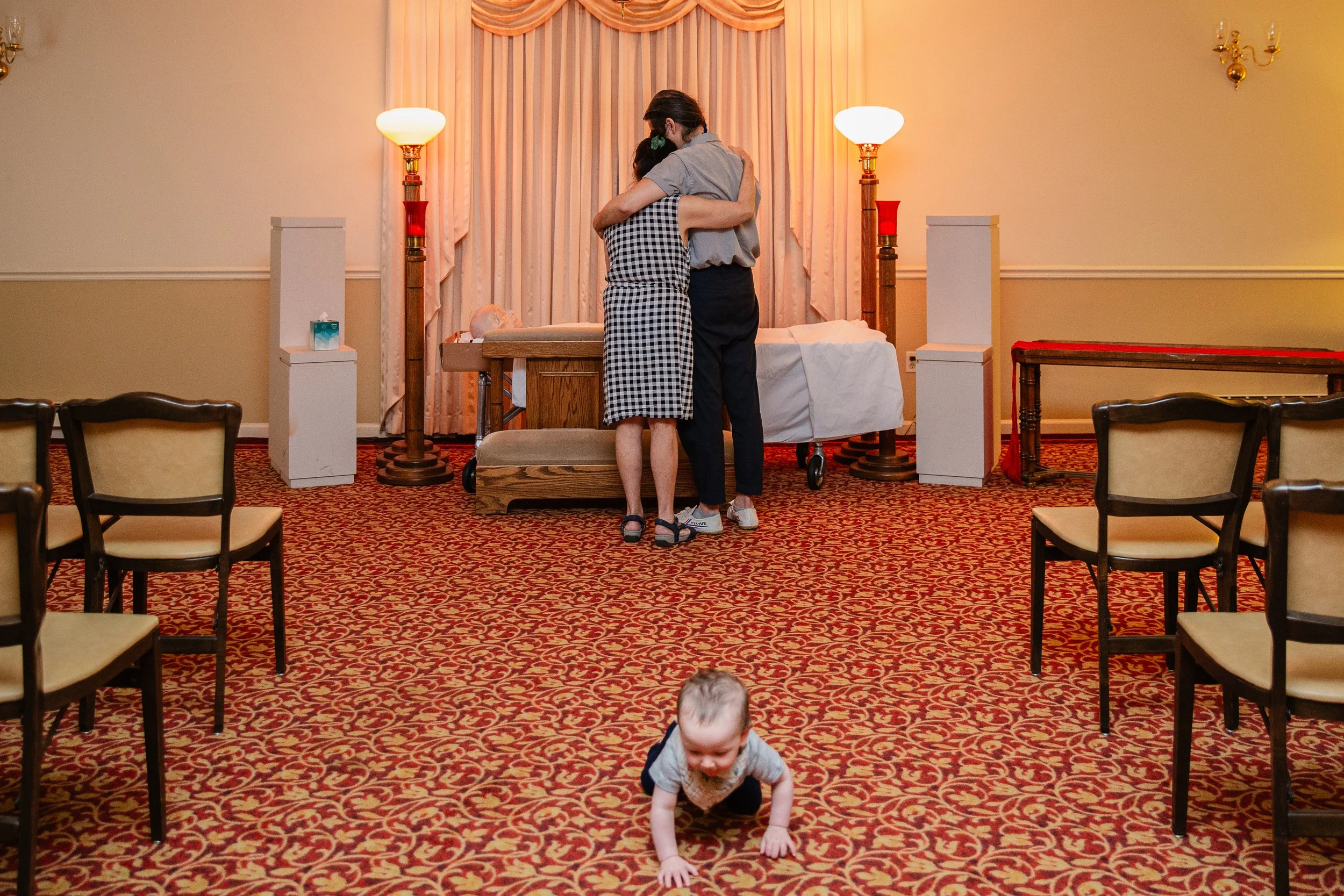 Two people hugging on a bed in a warmly lit room with pink curtains, while a crawling baby in the foreground explores the patterned carpet.