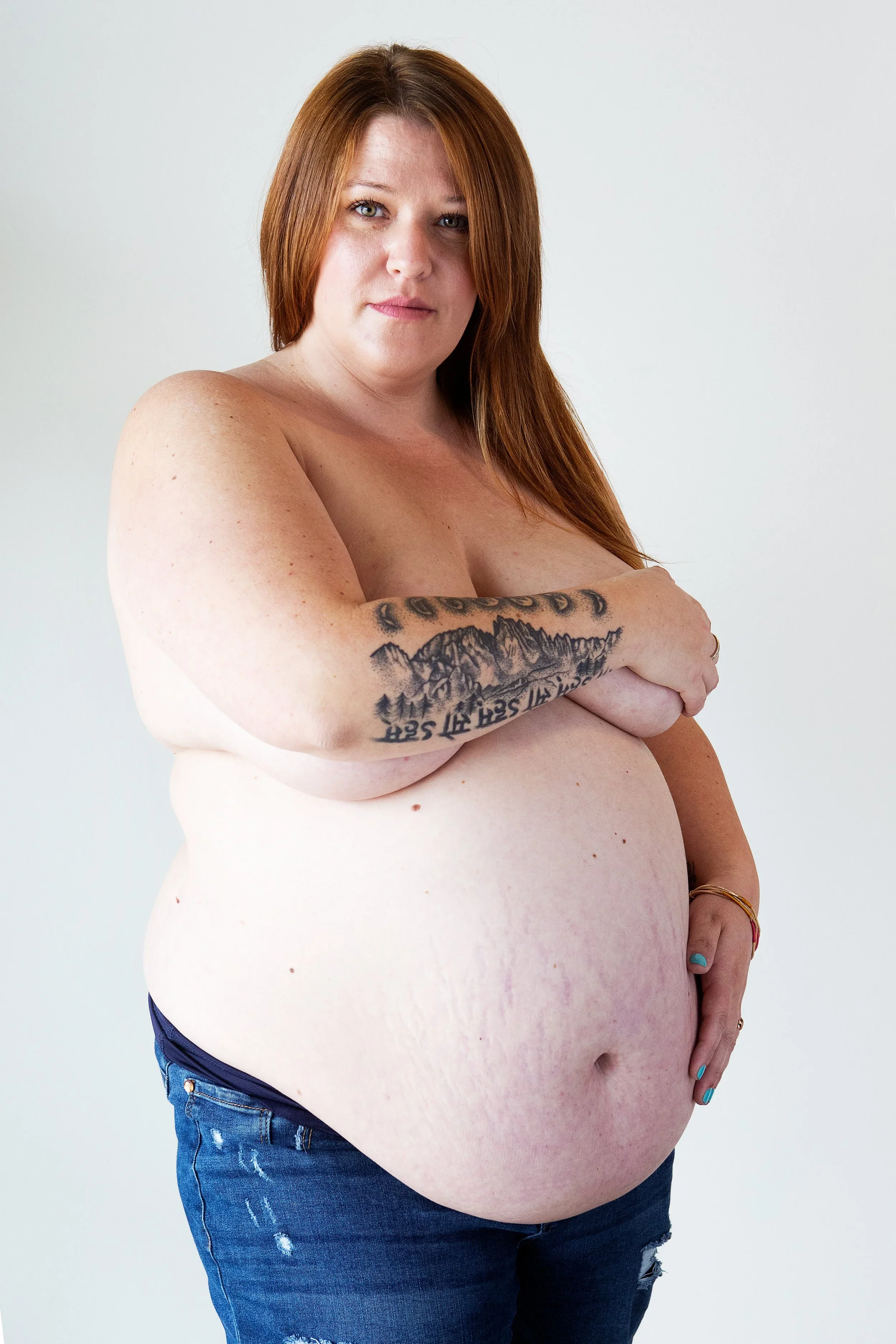 A topless woman with long red hair, blue jeans, and tattoos on her arm, standing against a plain white background.