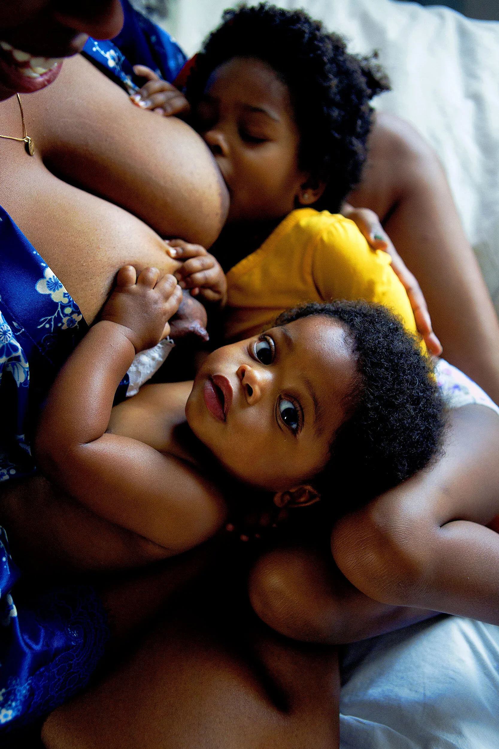 A woman breastfeeding two children, one child is breastfeeding while the other child is lying beside her.