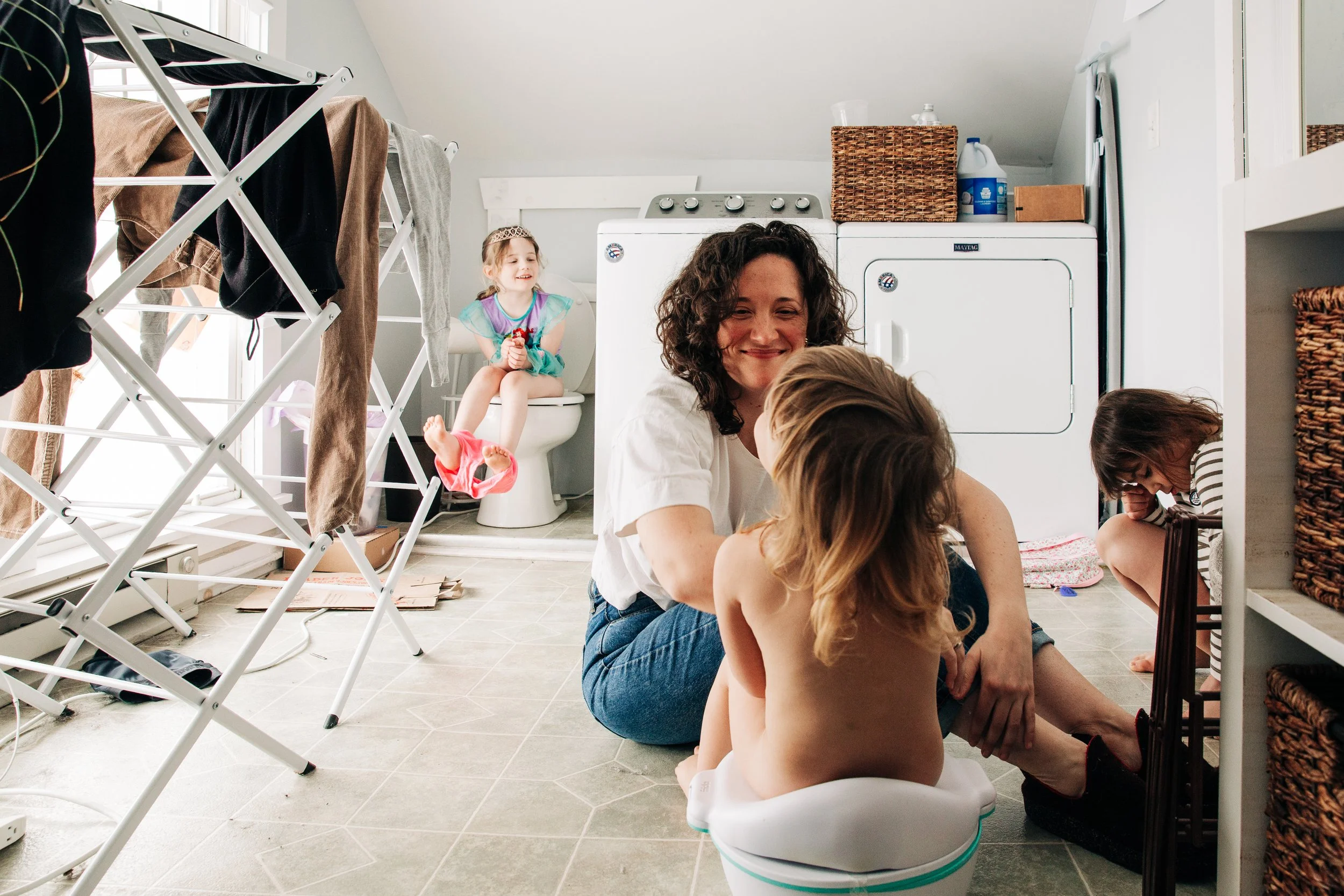 A woman and four children playing on the bathroom floor next to a washing machine and dryer, with laundry drying racks and a toilet in the background.