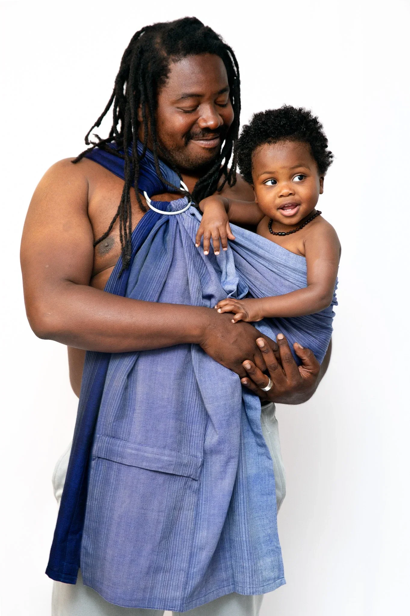 An adult man with long dreadlocks and a goatee, smiling with his eyes closed, holding a young child in a blue wrap carrier. The child has short curly hair, wearing a black beaded necklace and smiling, against a plain white background.