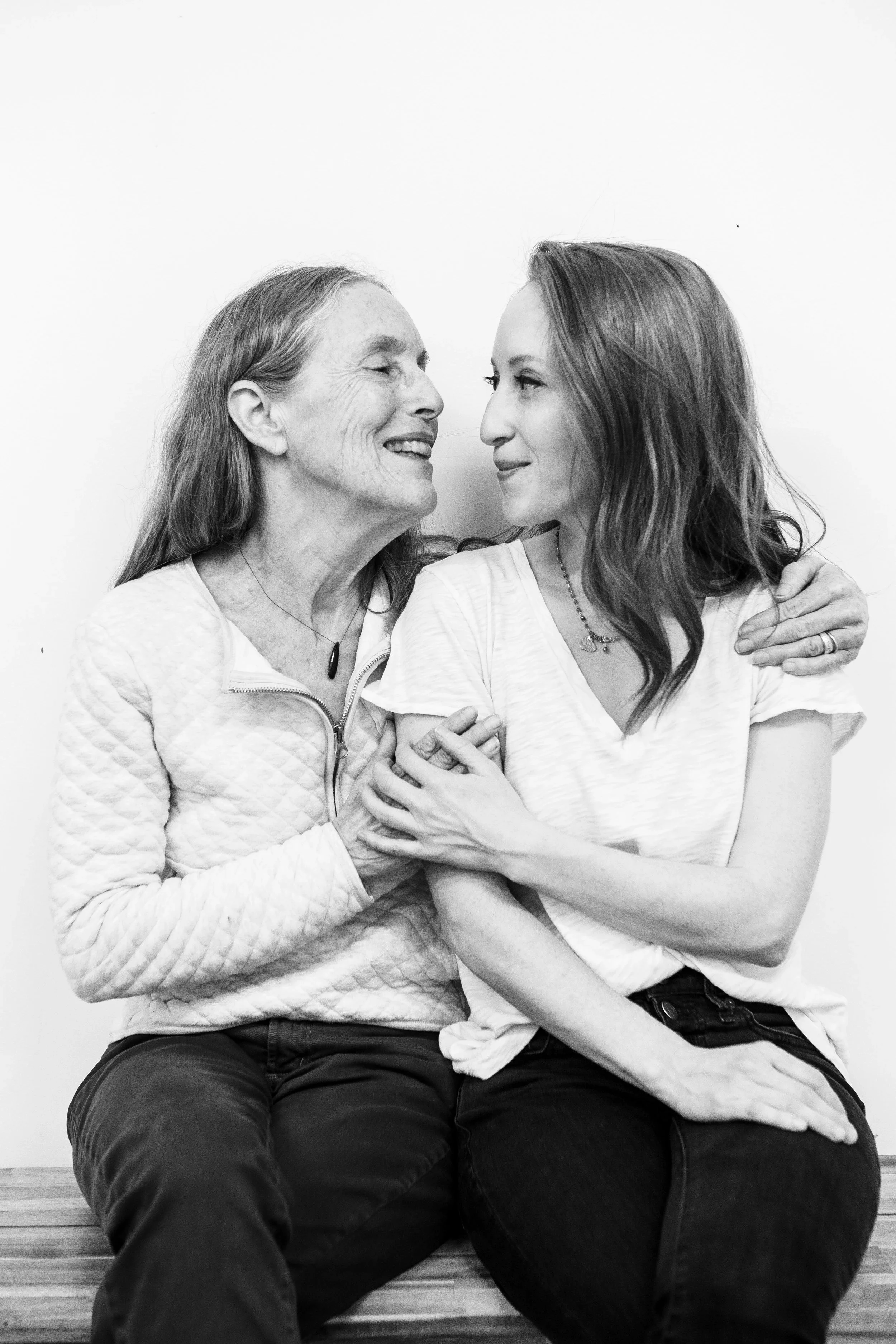 A black and white photo of an elderly woman and a younger woman sitting closely together on a bench, smiling at each other with their faces inches apart. The elderly woman has long hair and is wearing a quilted jacket, while the younger woman has sho