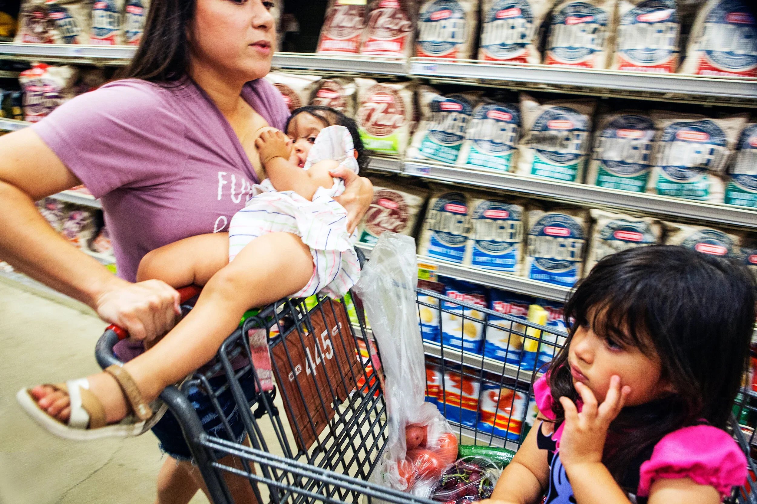 A woman breastfeeding a baby in a shopping cart while shopping in a grocery store, with two children nearby and shelves stocked with bags of flour in the background.