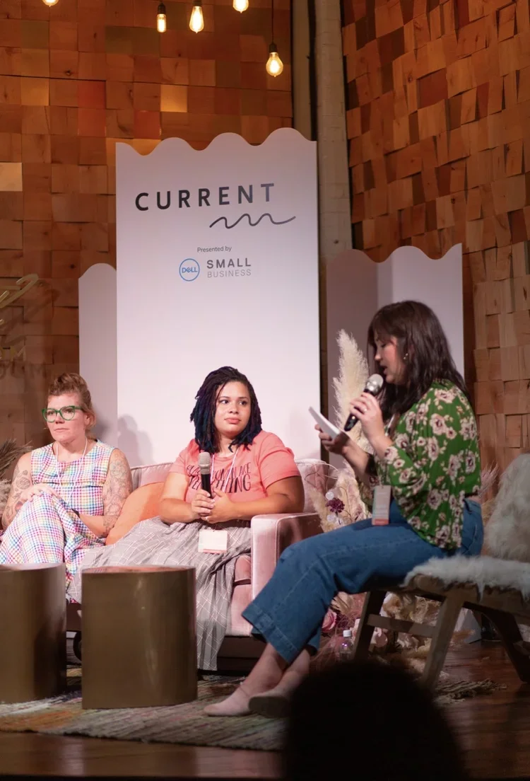 Three women sitting on a stage during a panel discussion at an event called 'Current,' presented by Dell Small Business, with one woman speaking into a microphone and the other two listening. The stage has warm lighting and wooden decor.