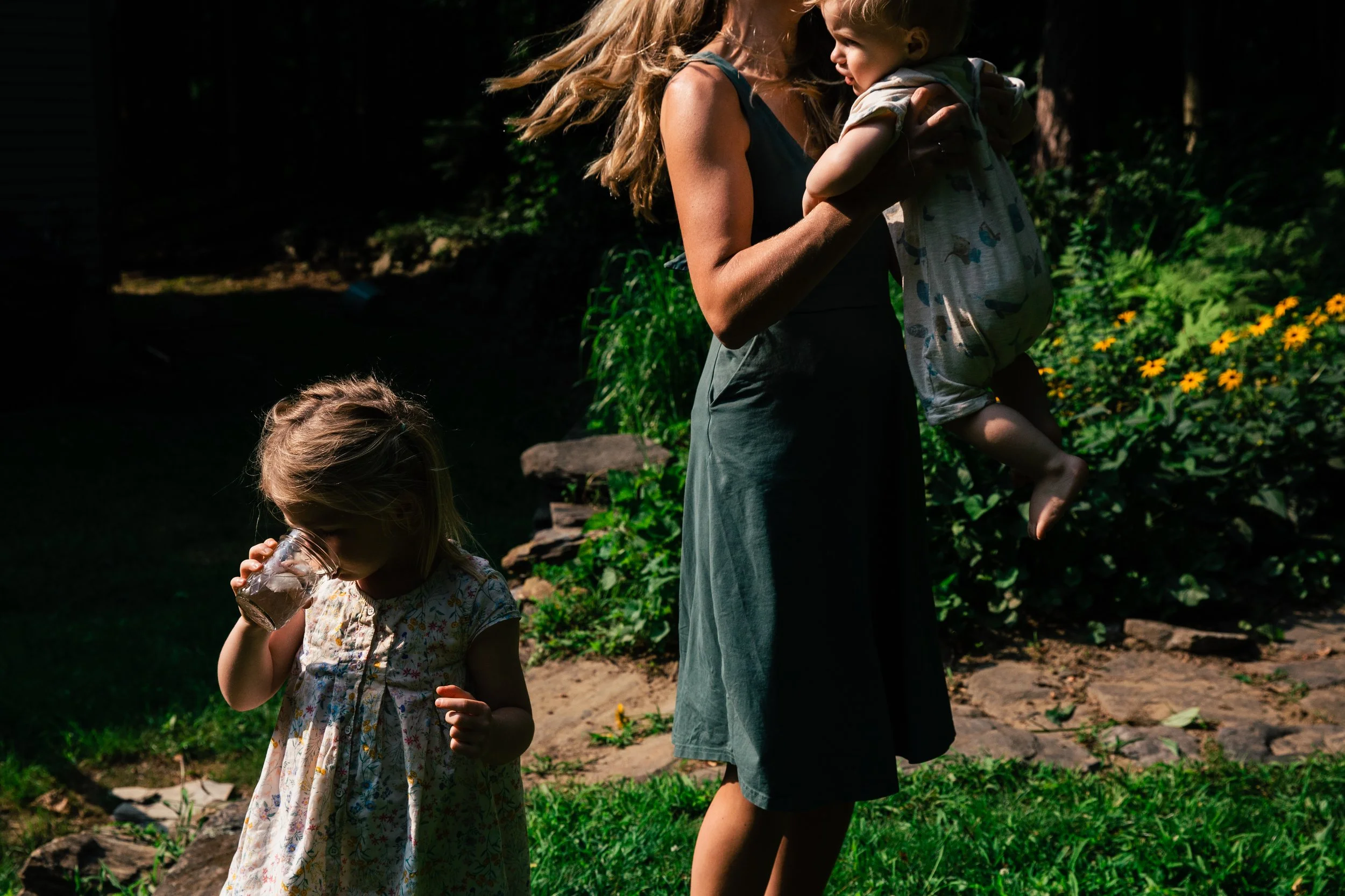 A woman holding a young girl in a garden with green plants and yellow flowers, while another girl drinks from a glass.