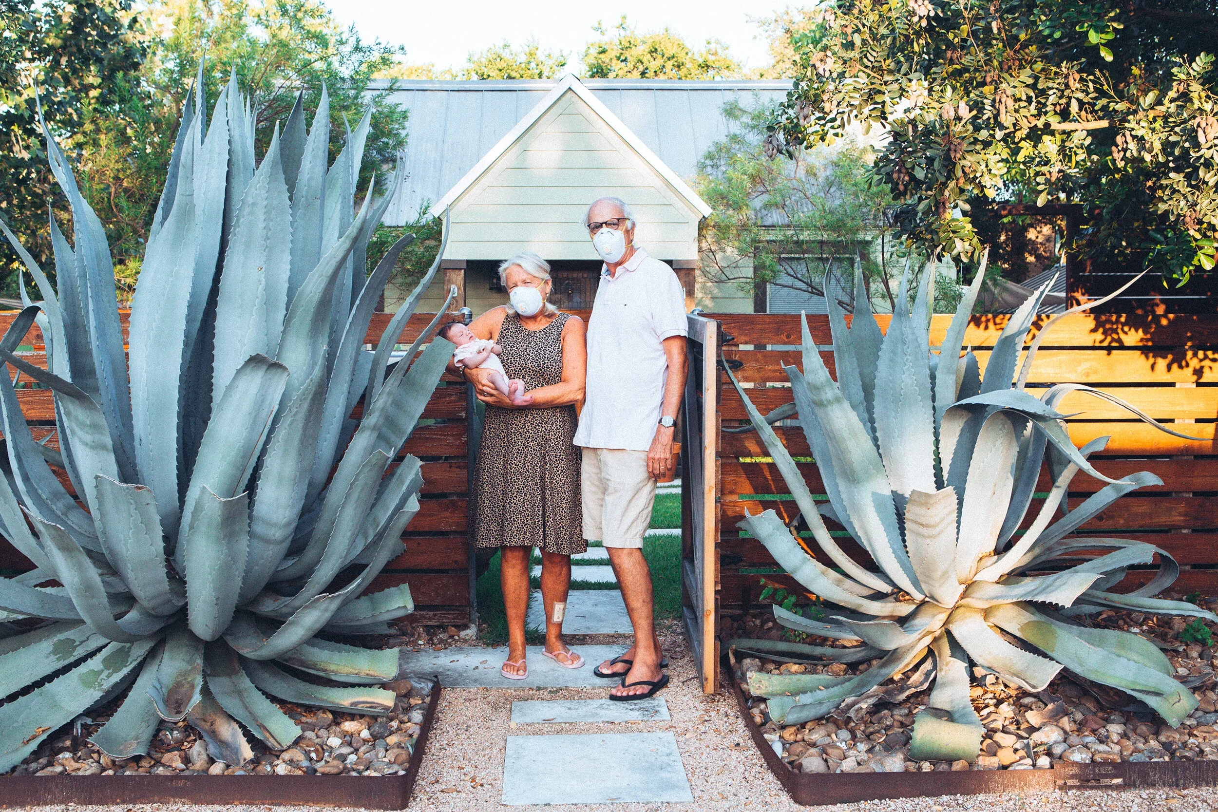 Three older adults wearing face masks stand behind a large agave plant in a backyard garden with a white house in the background.
