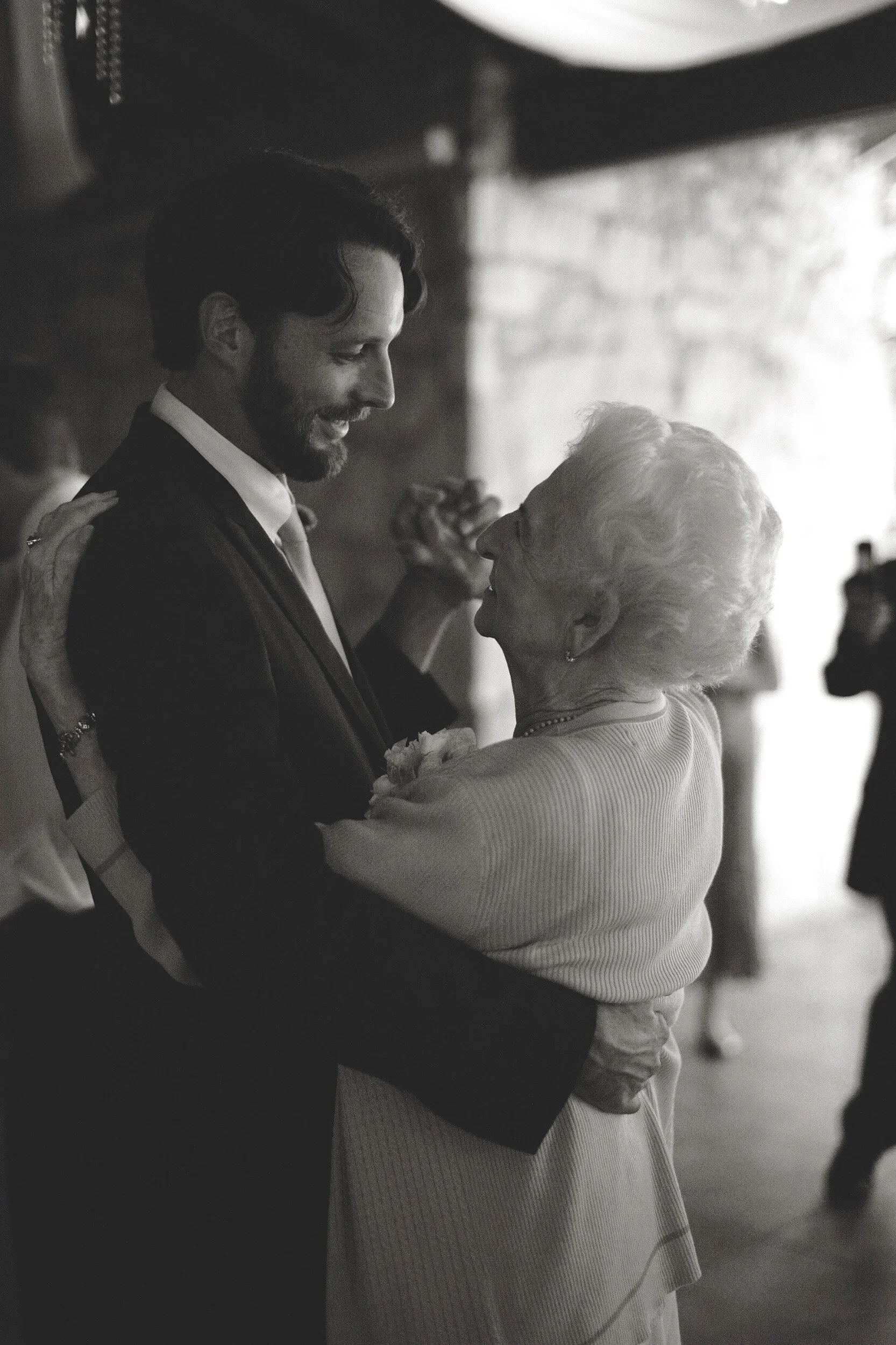 A young man with dark, wavy hair and a beard dressed in a suit dancing with an elderly woman with short, curly hair and a pearl necklace, indoors with blurred background.