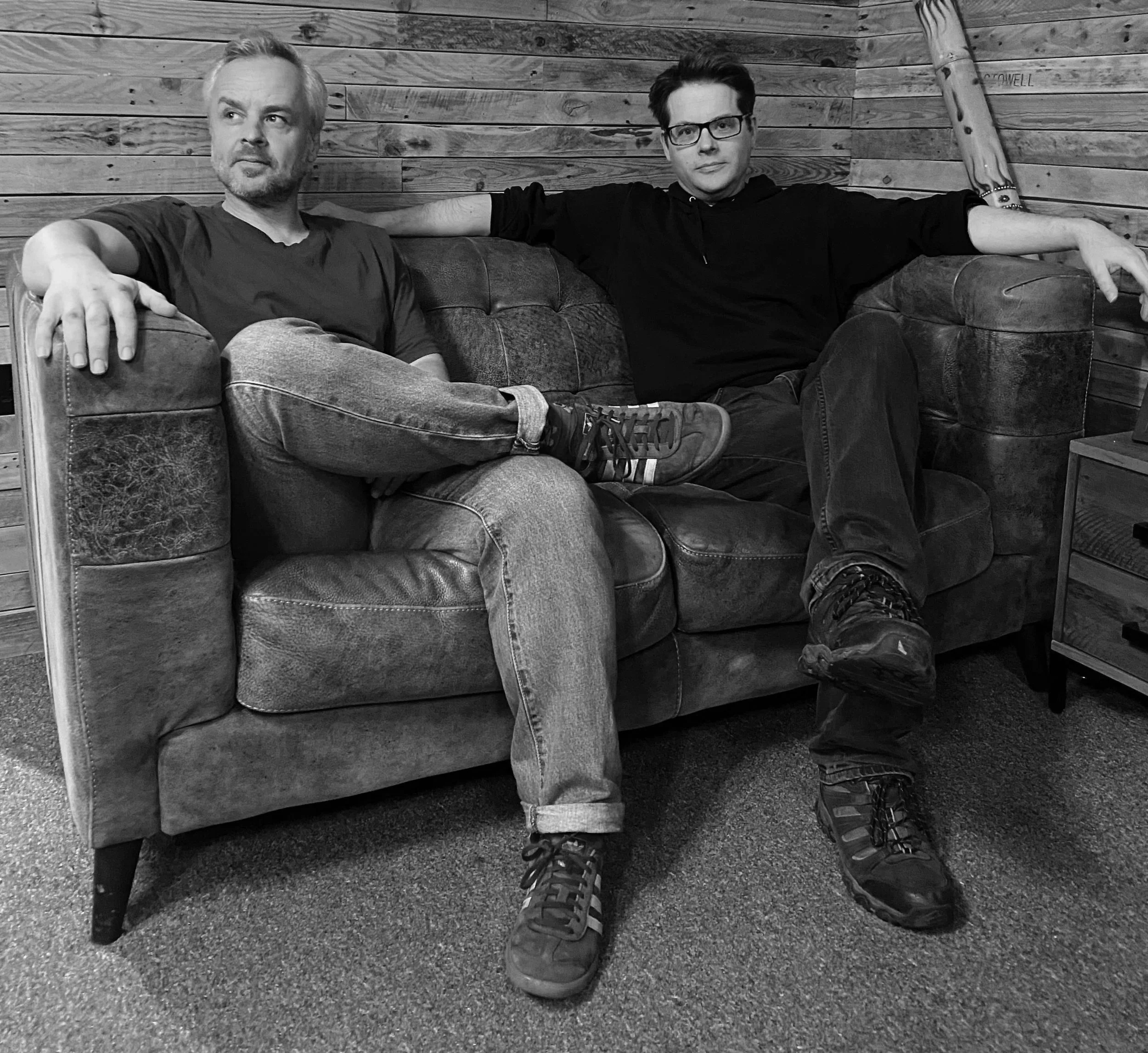 Two men sitting on a couch with wood wall background, one wearing glasses and black hoodie, the other with gray hair and beard, both casually dressed. Image is in black and white.