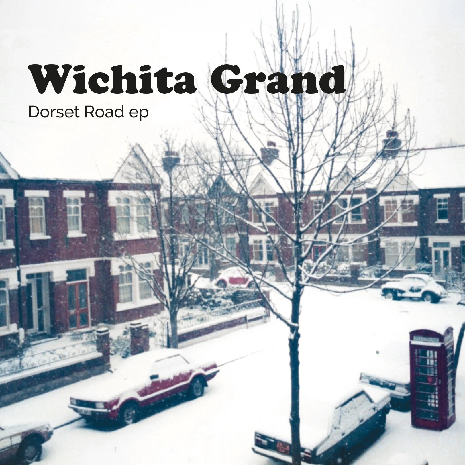 Snow-covered street scene with parked cars, a brick apartment building, a snow-covered telephone booth, and leafless trees during winter daytime.