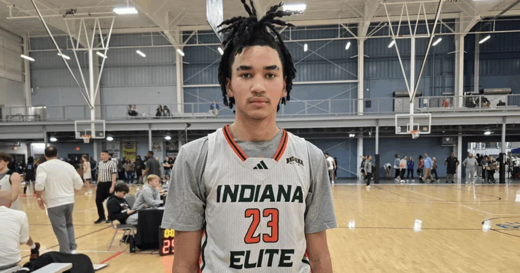 A young man with dreadlocks standing on a basketball court wearing a gray Indiana Elite basketball jersey with the number 23. The gym has a high ceiling with metal beams and basketball hoops, with people walking and sitting in the background.