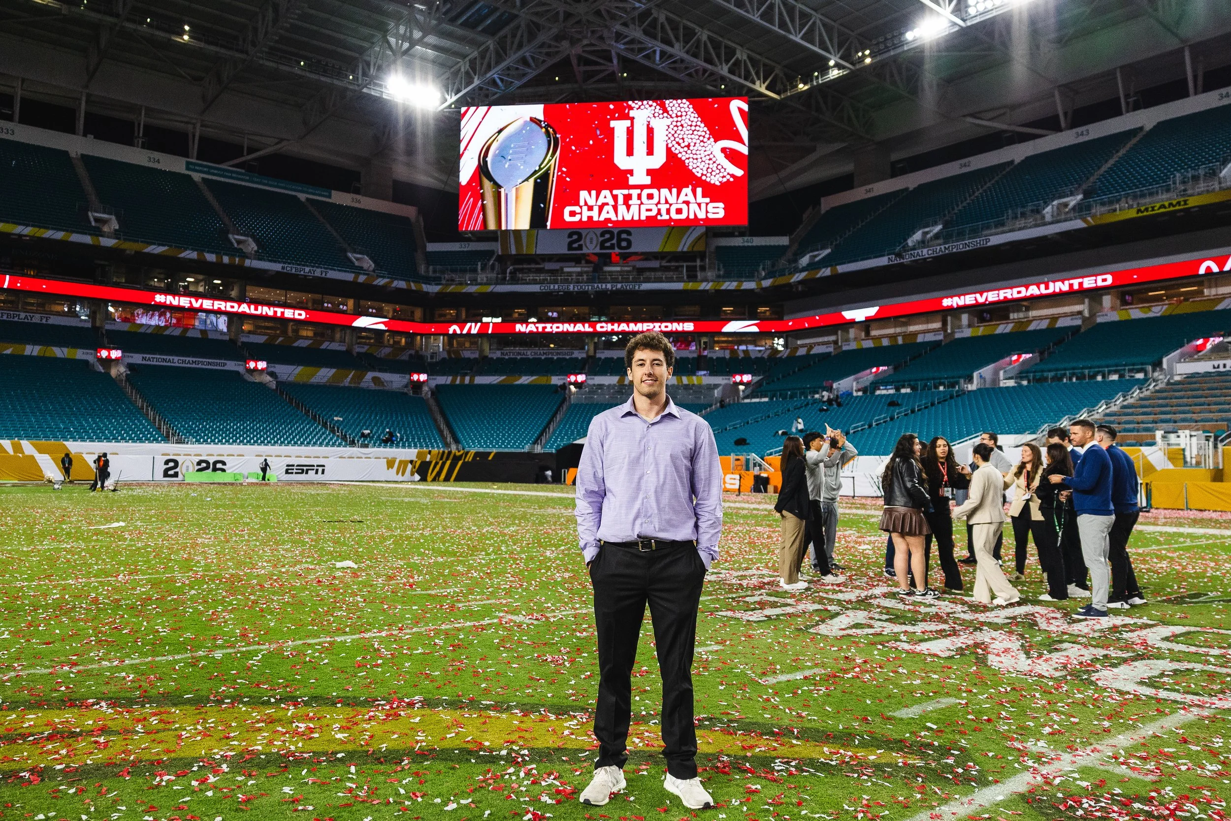 A man in a light purple shirt and black pants standing on a football field decorated with red and white confetti, inside a large empty stadium with a large screen displaying 'National Champions' and a trophy, with people gathered in the background after a sporting event celebration.