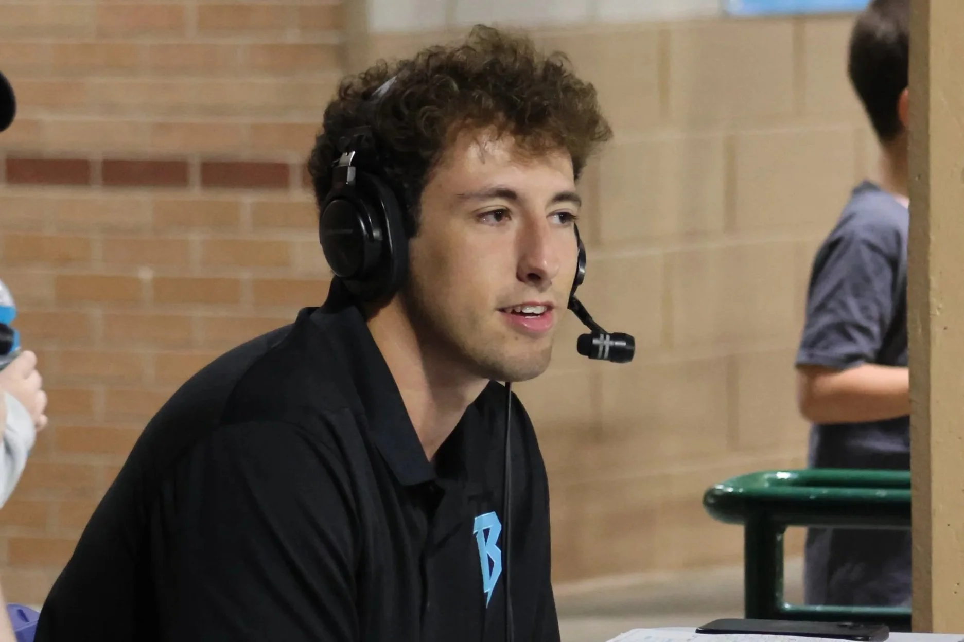 A young man with curly brown hair wearing a black shirt and headset with microphone, sitting indoors against a brick wall.
