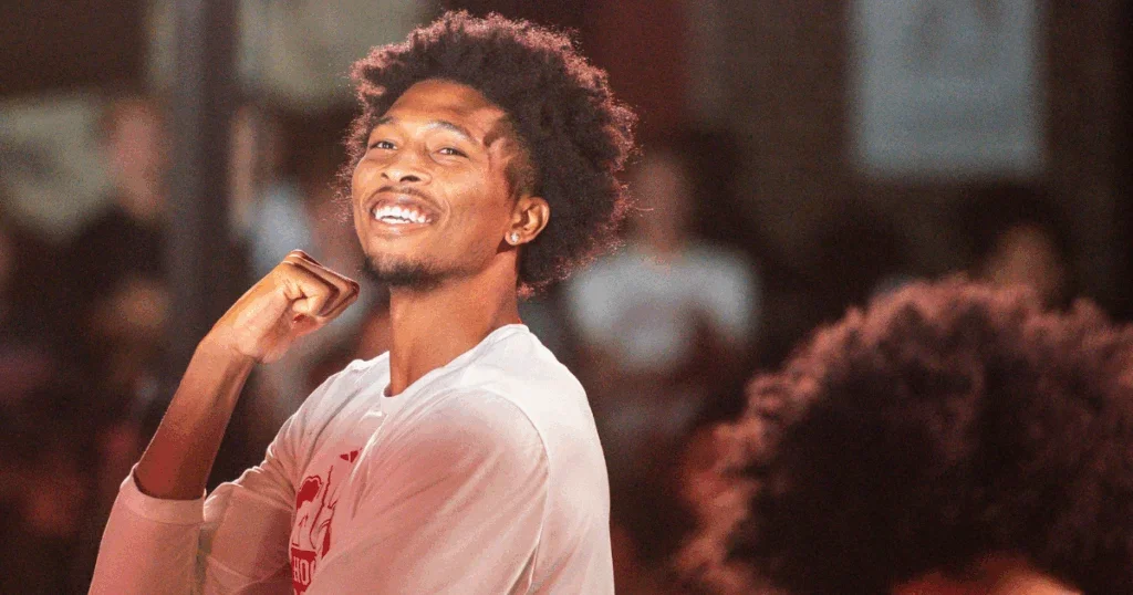 A young man with curly hair flexing his arm and smiling, in a crowded indoor setting.