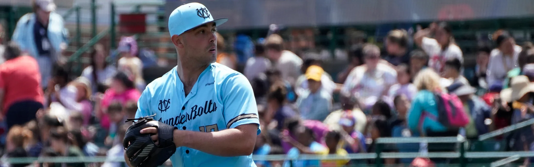 A male baseball player wearing a light blue uniform and cap, standing on the field with a glove on his left hand, and a crowd of spectators in the background.