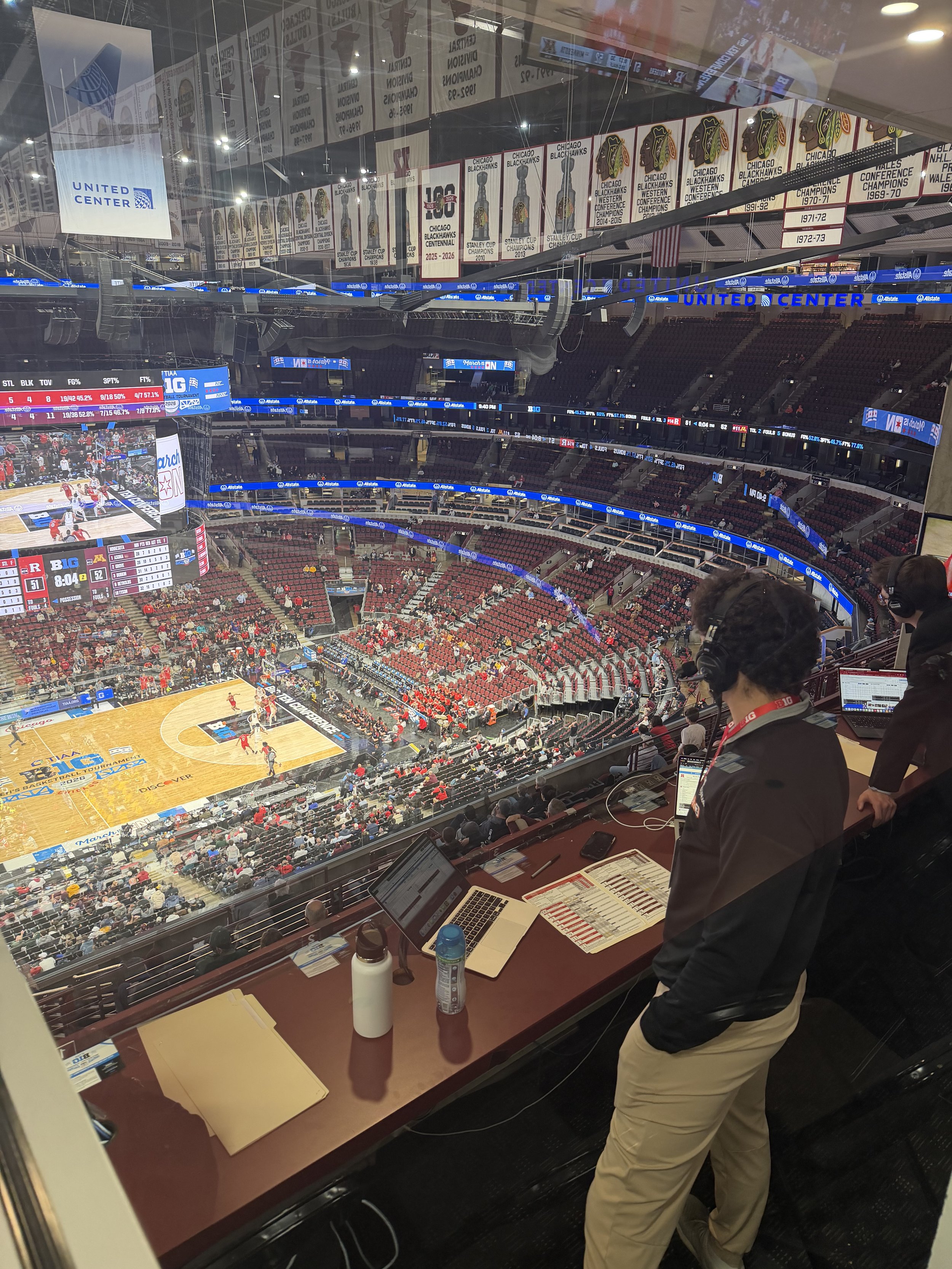 The interior of a sports arena with players on the basketball court during a game, and spectators seated in the stands. A press box area with computers, water bottles, and people working is visible in the foreground.