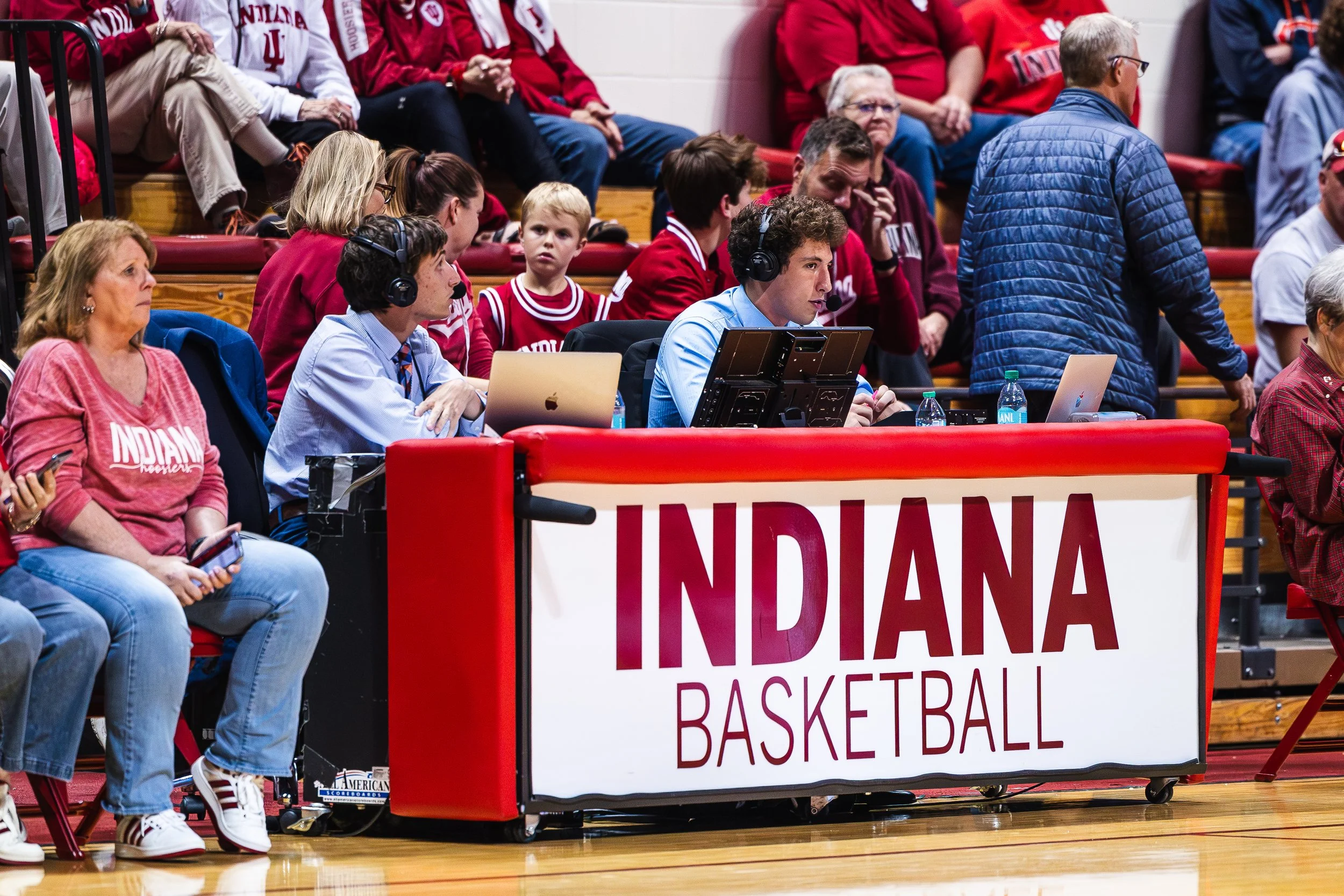 People sitting courtside at a basketball game, some wearing Indiana University apparel, with a sign reading "INDIANA BASKETBALL." A man and woman with headsets and laptops are seated at a broadcasting table.