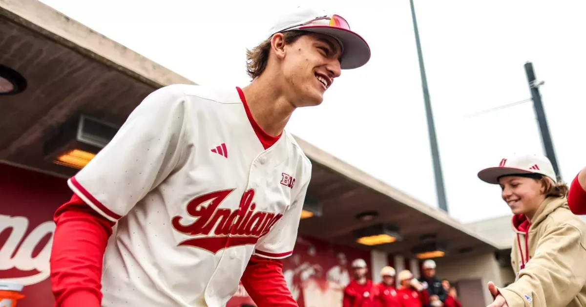 A young man in an Indiana baseball jersey and cap smiling at the dugout, with a woman in a hoodie and cap smiling nearby, in a baseball stadium.