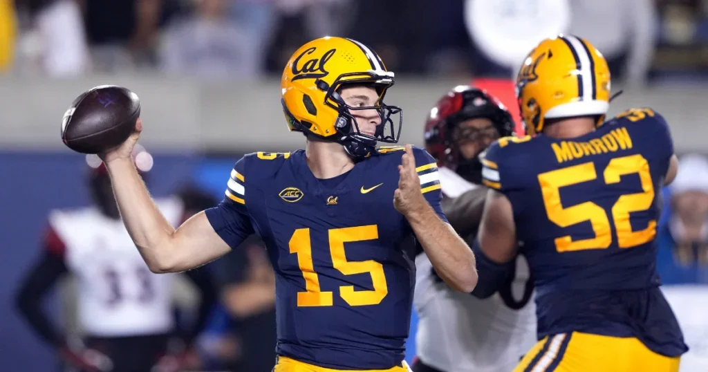 A college football player wearing a navy and yellow uniform with the number 15, holding a football in his right hand, appears to be preparing to throw a pass. The helmet has 'Cal' written on it, indicating University of California. Another player wearing jersey number 52 is visible in the background.