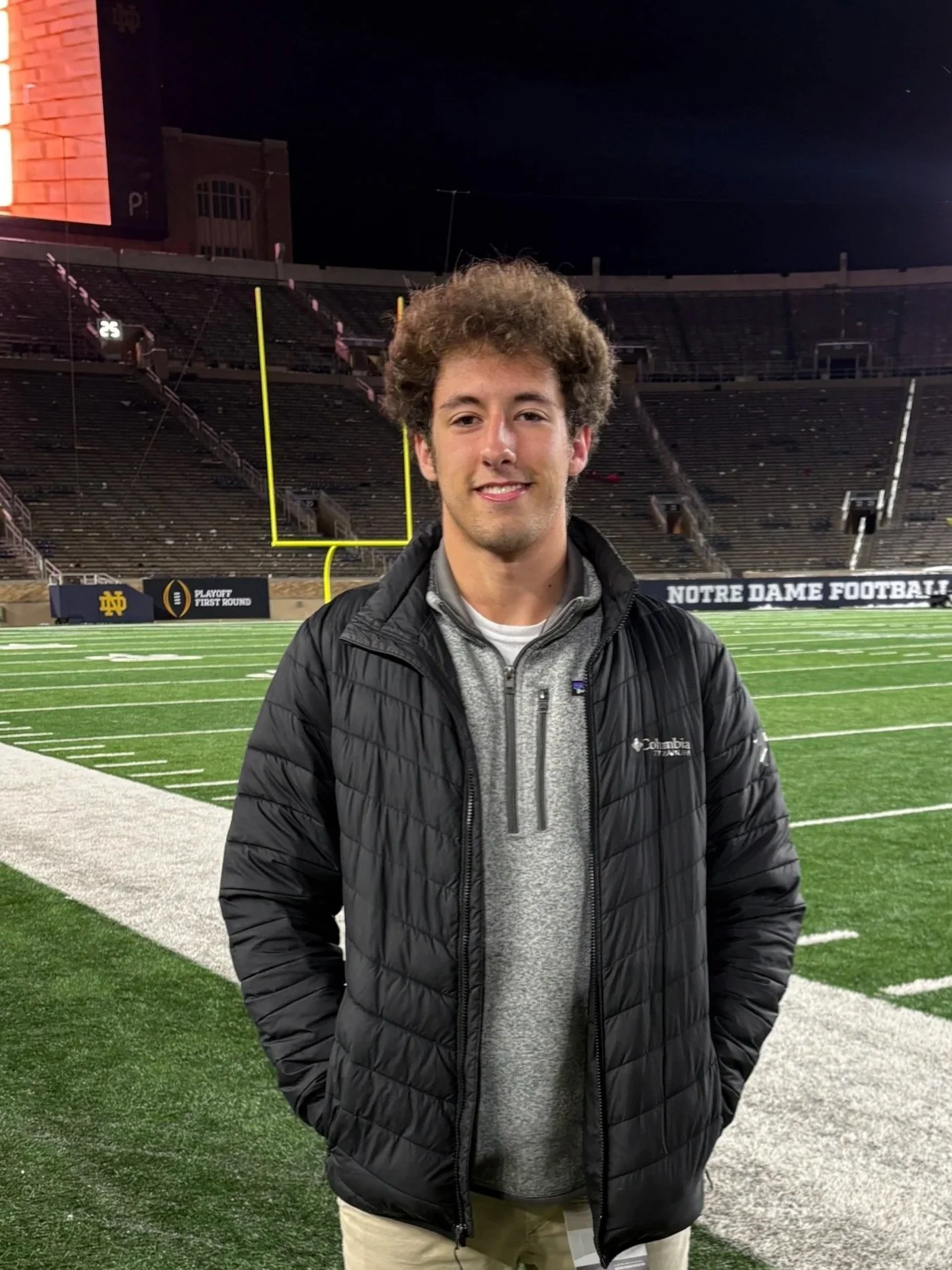 Young man standing on a football field at night, wearing a gray hoodie and black puffer jacket, with empty stadium seats and goalposts in the background.
