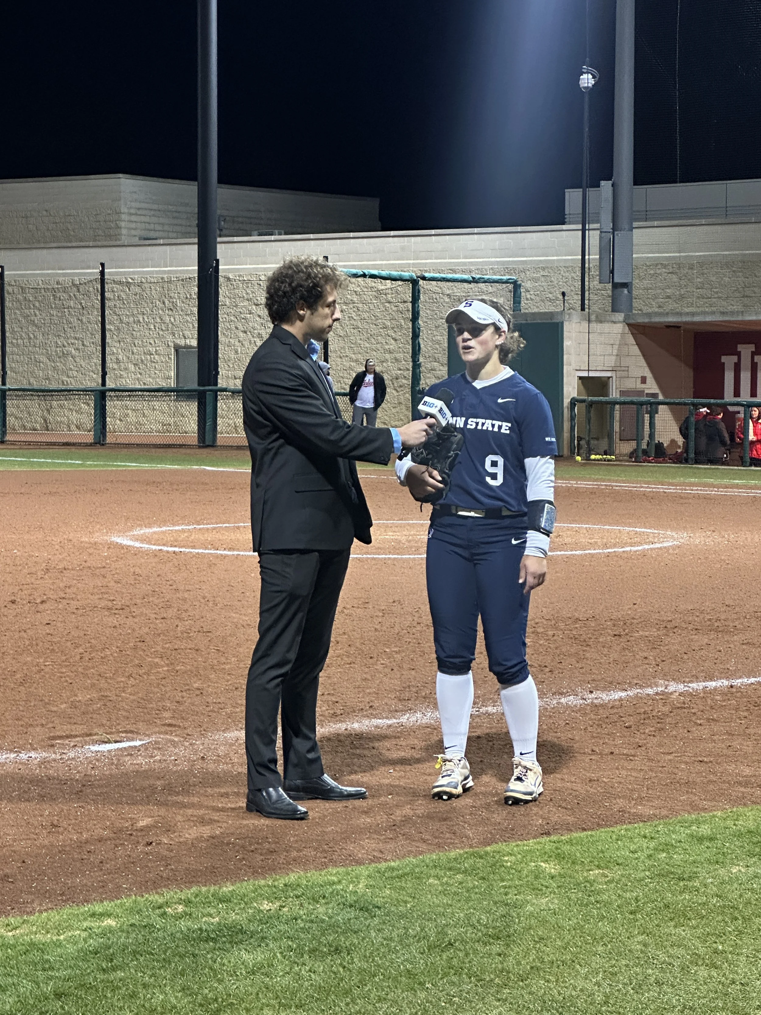 A woman in a softball uniform being interviewed by a man holding a microphone on a baseball field at night. The woman is wearing a blue jersey with the number 9, white pants, and a white cap. There are a few people in the background near the fence.