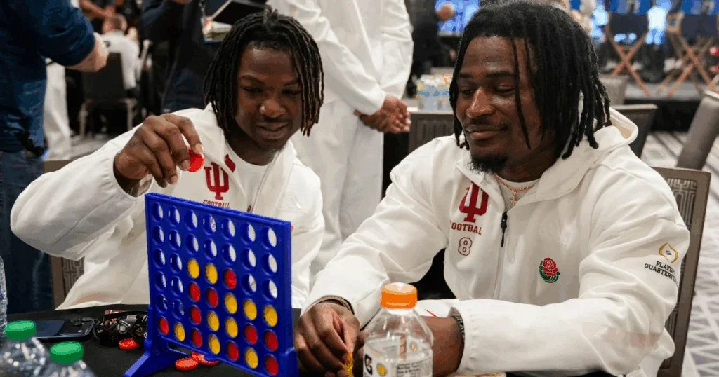 Two men in white Indiana football jackets playing Connect Four at a table, one placing a red disc into the game while the other watches, with a crowd in the background.
