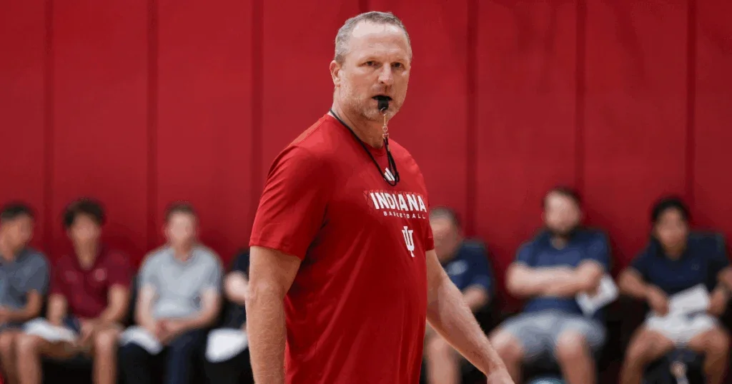A volleyball coach in a red Indiana basketball shirt on a court during a game or practice, with players seated in the background.
