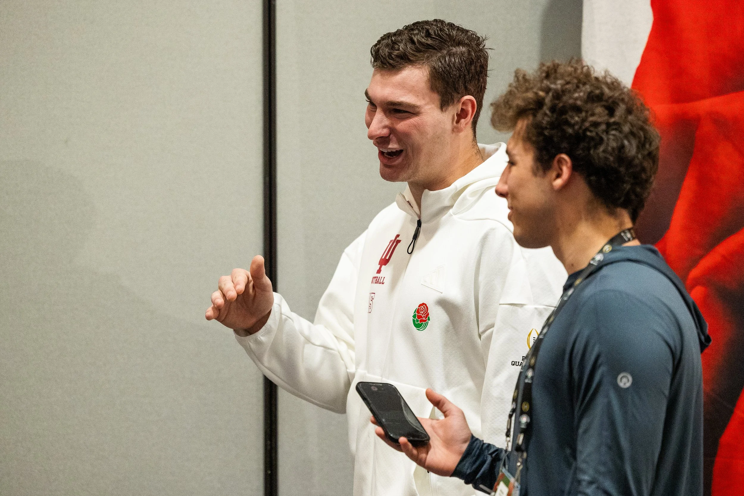 Two young men are sitting and talking, one wearing a white jacket with a red emblem, and the other in a dark jacket with a phone in hand. They are indoors against a plain grey wall, possibly at a sports or academic event.