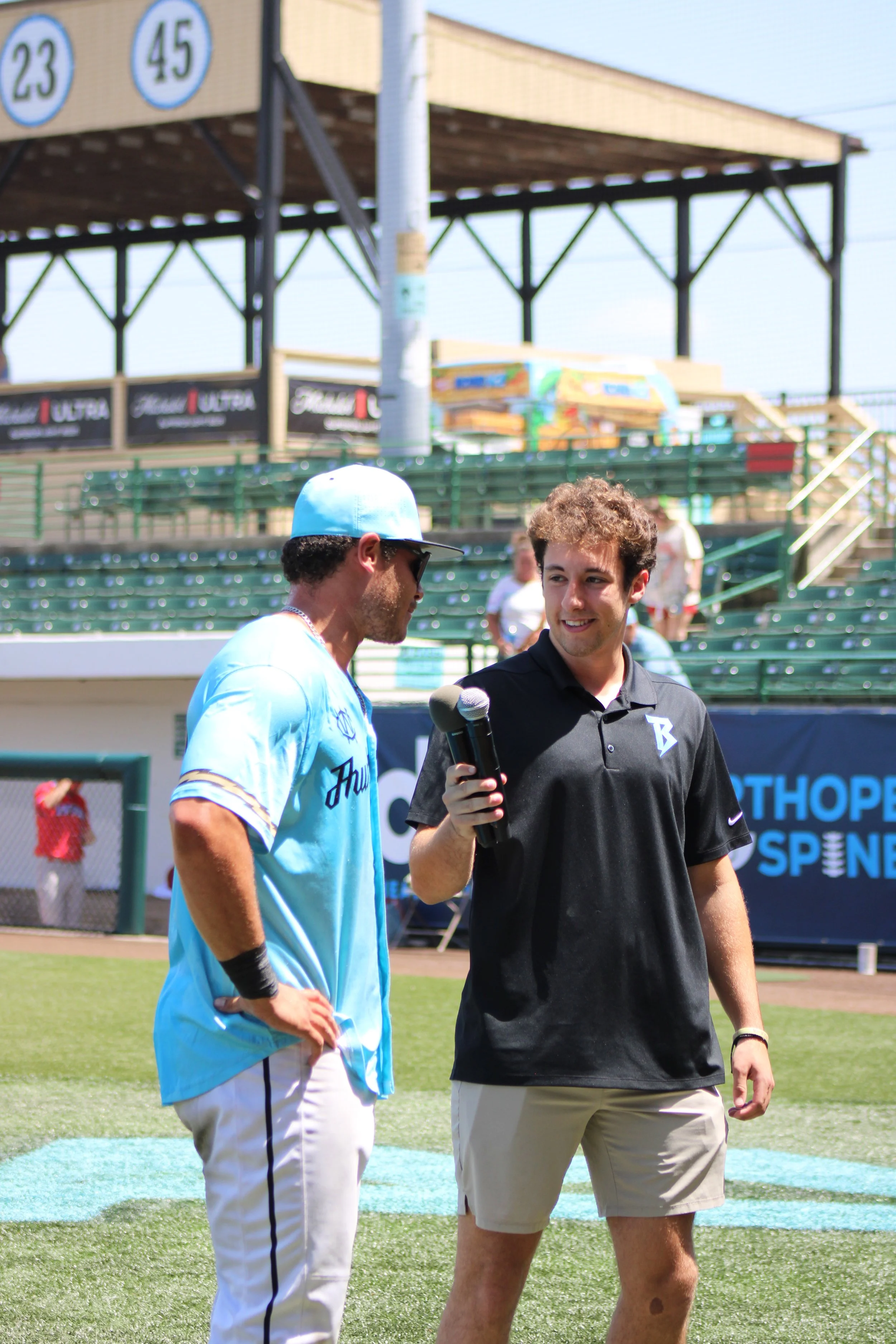 Two young men are standing on a baseball field, engaged in conversation. One is wearing a light blue baseball jersey and a matching cap, while the other is dressed in a black polo shirt and beige shorts, holding a microphone. The background features bleachers, some spectators, and a structure with signs and advertisements.