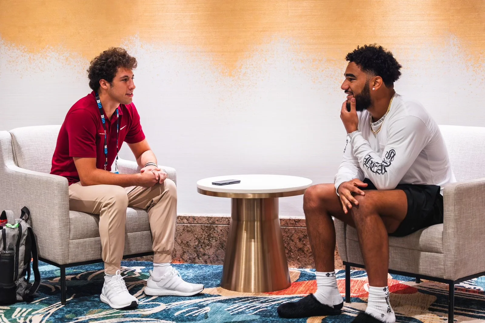 Two young men sitting on separate sofas facing each other, engaged in conversation. A small round table with a smartphone on it is between them. The setting appears to be a modern indoor space with a colorful carpet and a plain wall backdrop.