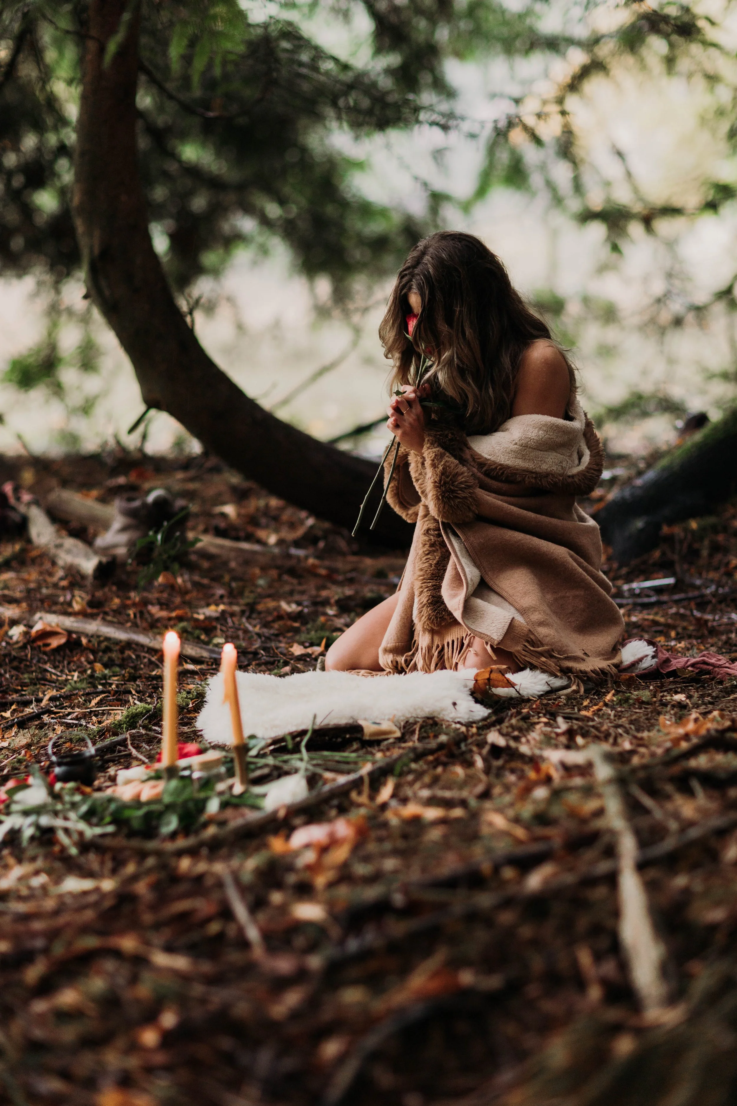 A woman with wavy brown hair kneeling in a forest, holding a flower close to her face, wrapped in a beige coat, surrounded by fallen leaves and a white cloth with candles in the foreground.