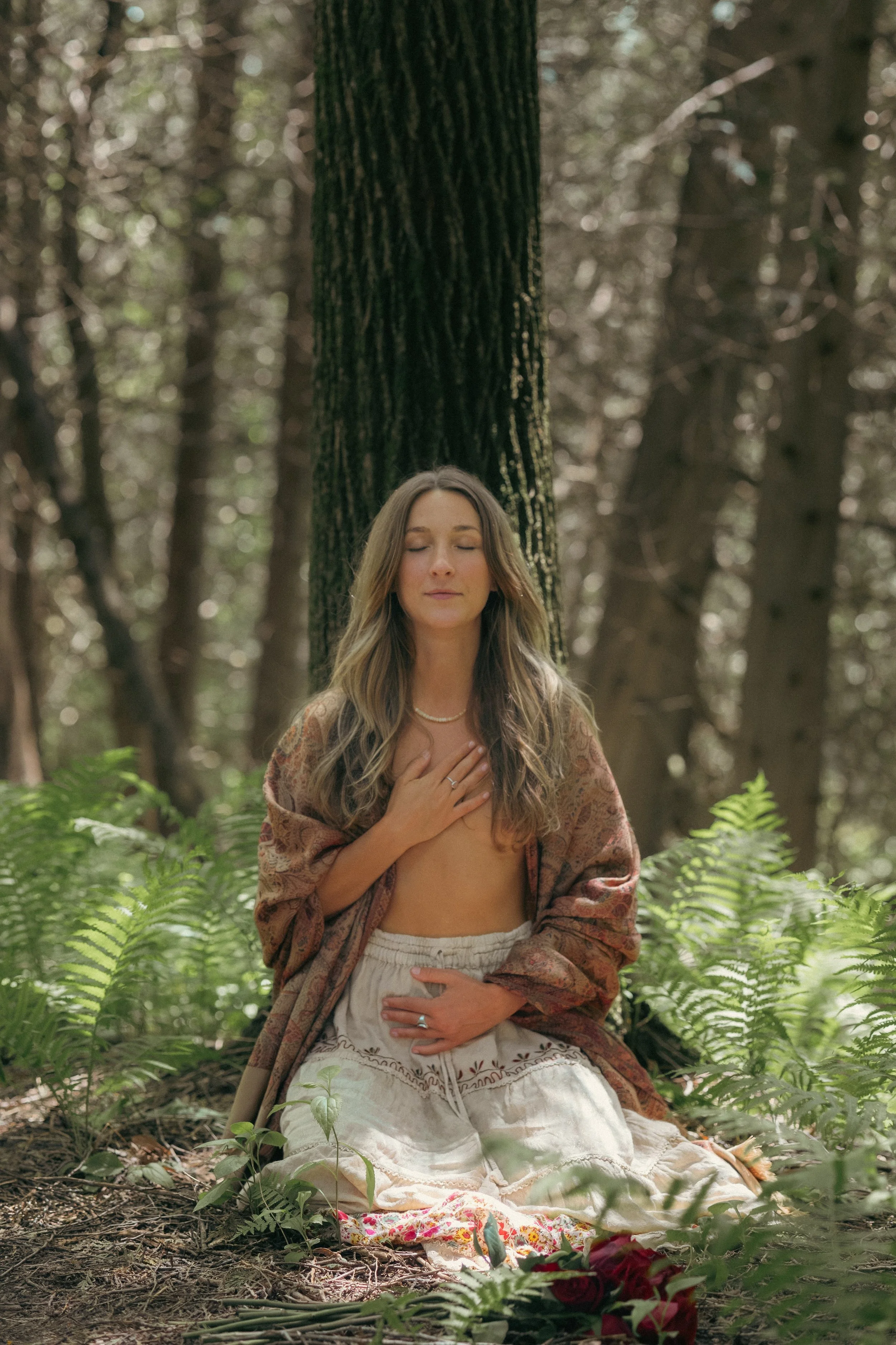 A woman with long wavy hair standing with her eyes closed, her right hand on her chest and left hand on her belly, in a forest setting. She is wearing a patterned shawl, white skirt, and has flowers and greenery at her feet.