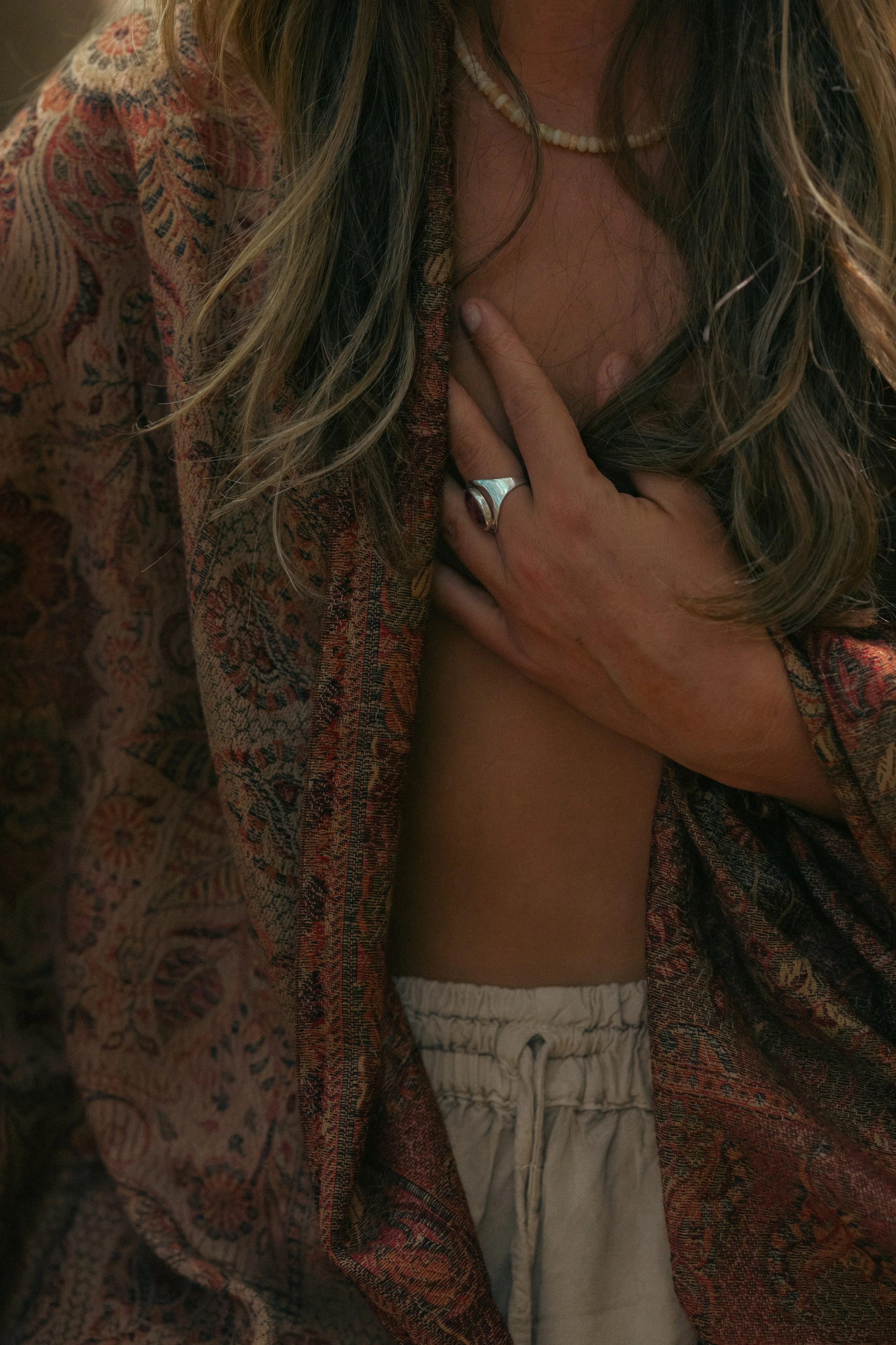Close-up of a woman wearing a patterned bohemian-style jacket, with long, wavy hair, a pearl necklace, and white pants, holding her chest with one hand.