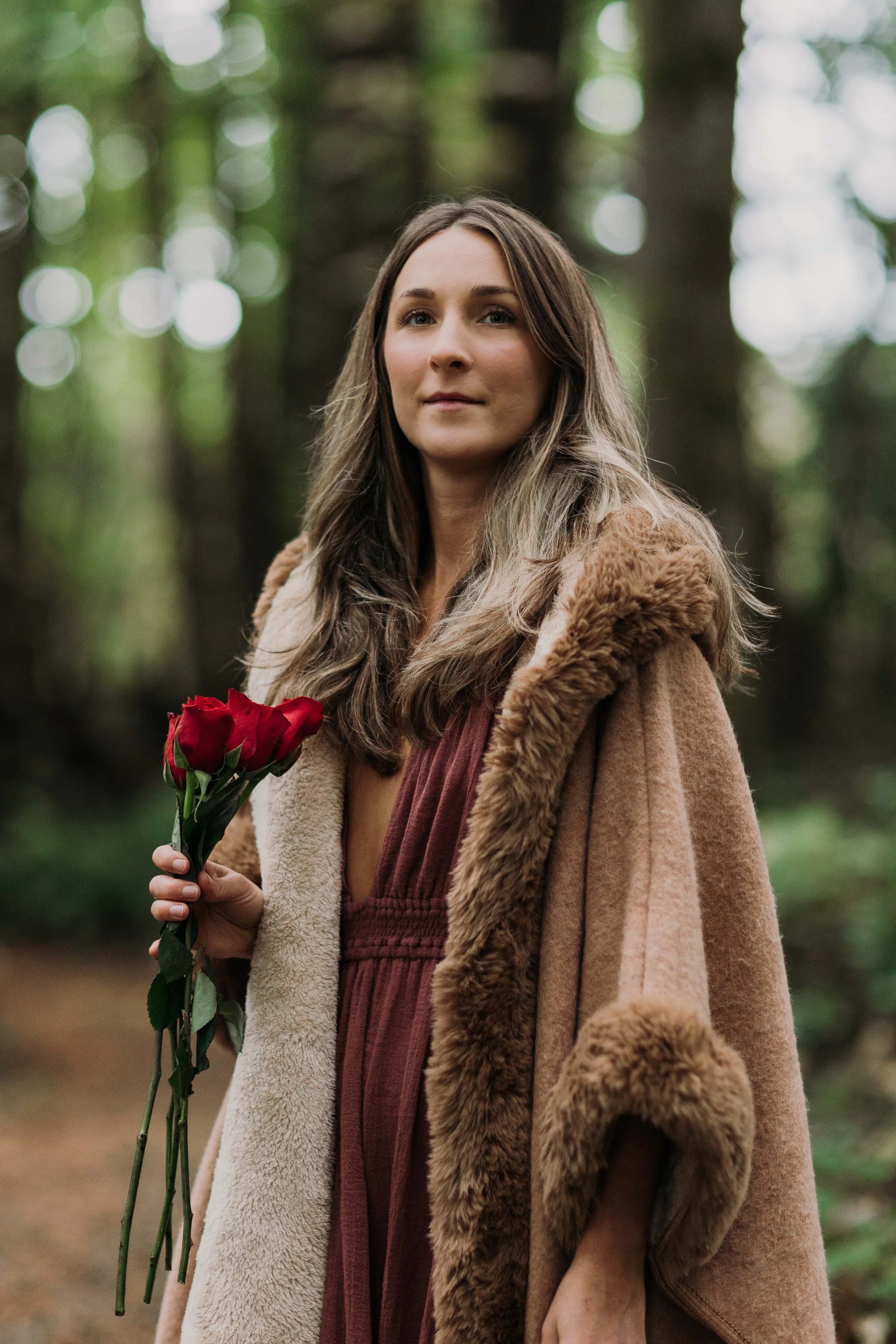 A woman with long brown hair holding a bouquet of red roses, wearing a long coat with her background of a forest with blurred trees and greenery.