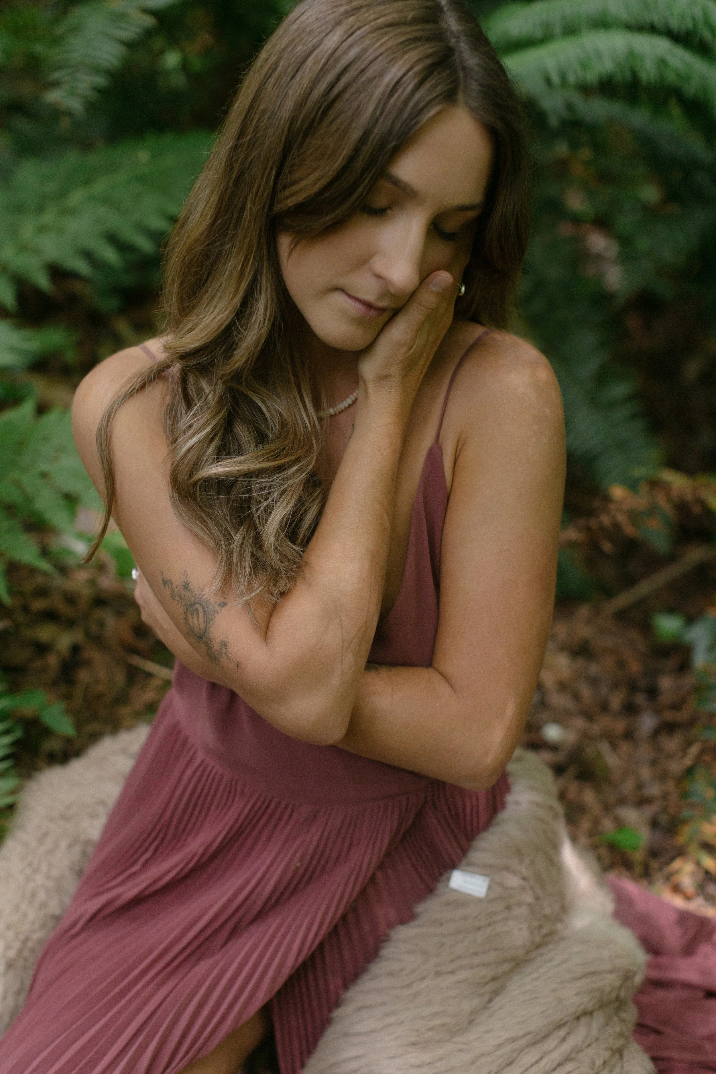 A woman with long, wavy brown hair and tattoos on her right arm, wearing a pink dress and a pearl necklace, sits with eyes closed and her hand on her cheek, in a forest setting.