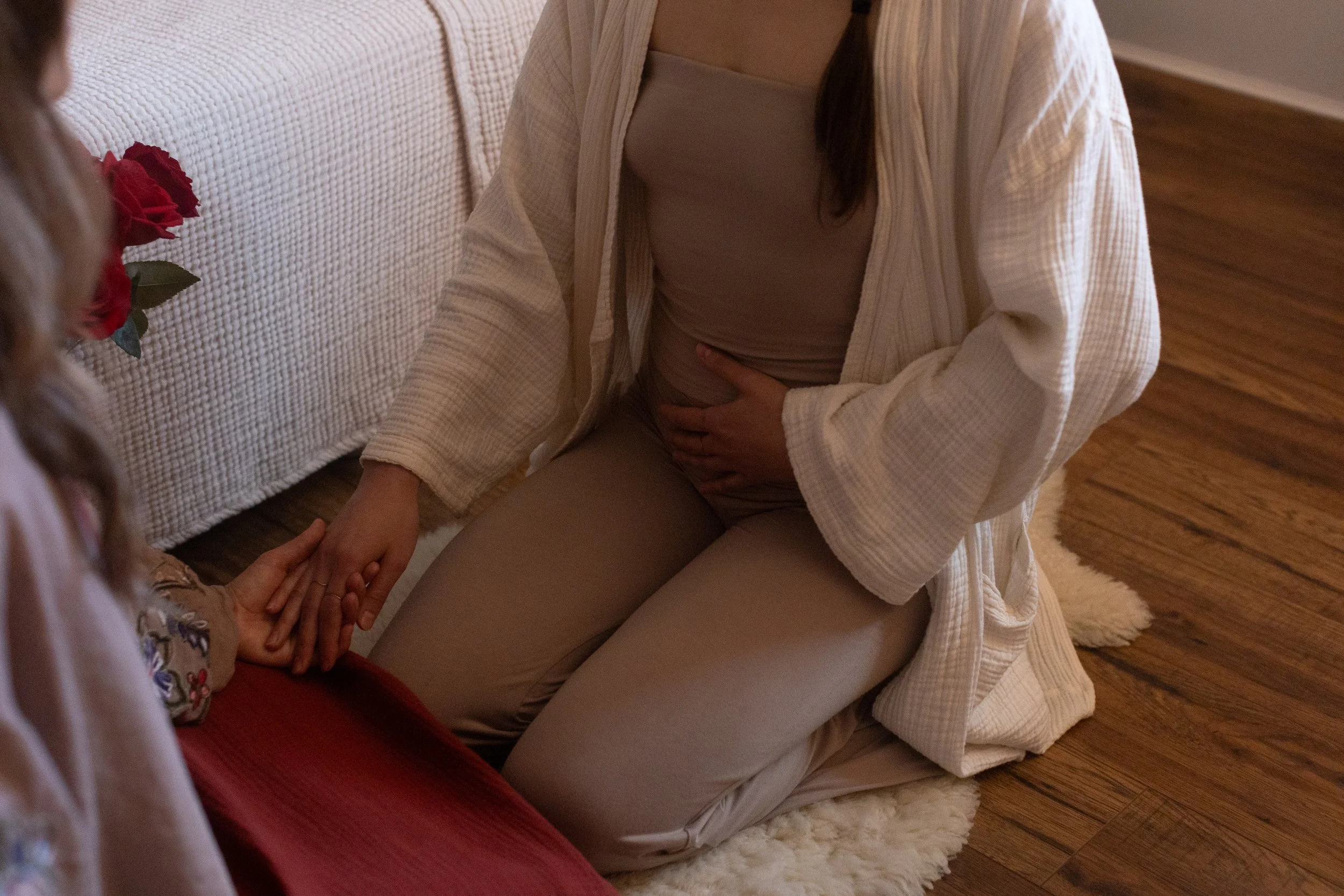 A woman kneeling on the floor next to a young girl, holding her hand in an embrace, inside a cozy room with hardwood floors, a rug, and a bed with a textured white bedspread and a red pillow.